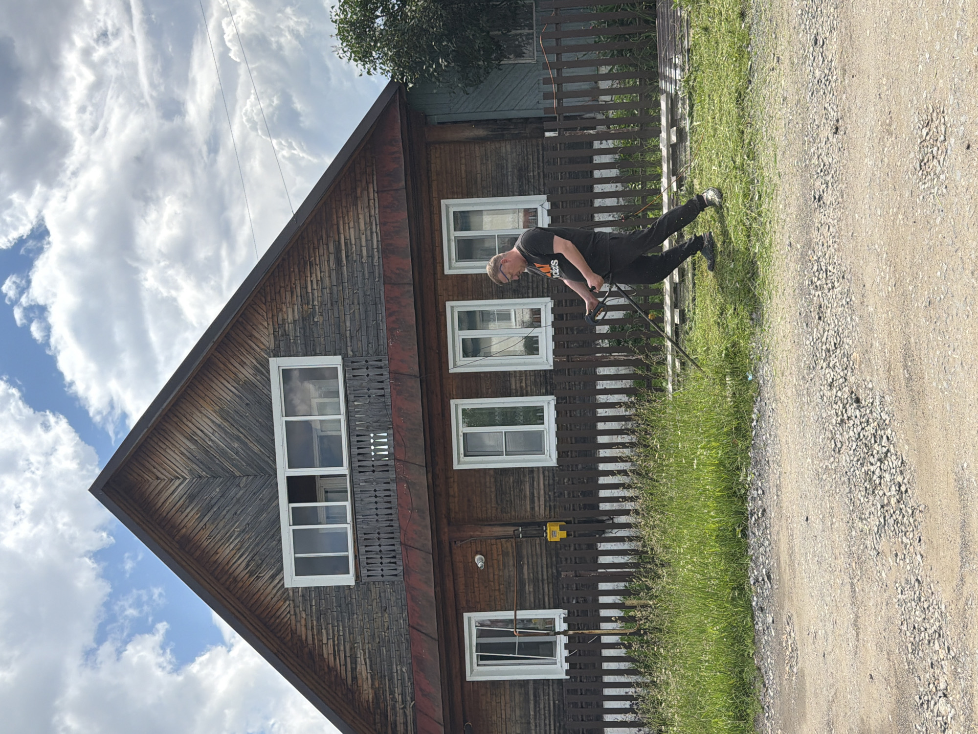 A man is mowing grass in front of a rustic wooden house in Mezhevoy, Russia. The man, wearing a black t-shirt and dark pants, is operating a string trimmer. He is positioned in the middle right of the frame, facing left, and appears to be mid-action. The house, which has a weathered wooden exterior and a steep gable roof, features several windows and a balcony. A wooden fence runs along the front of the property, separating the lawn from a gravel path in the foreground. The sky above is partly cloudy. The overall scene depicts a common domestic activity in a rural or semi-rural setting.