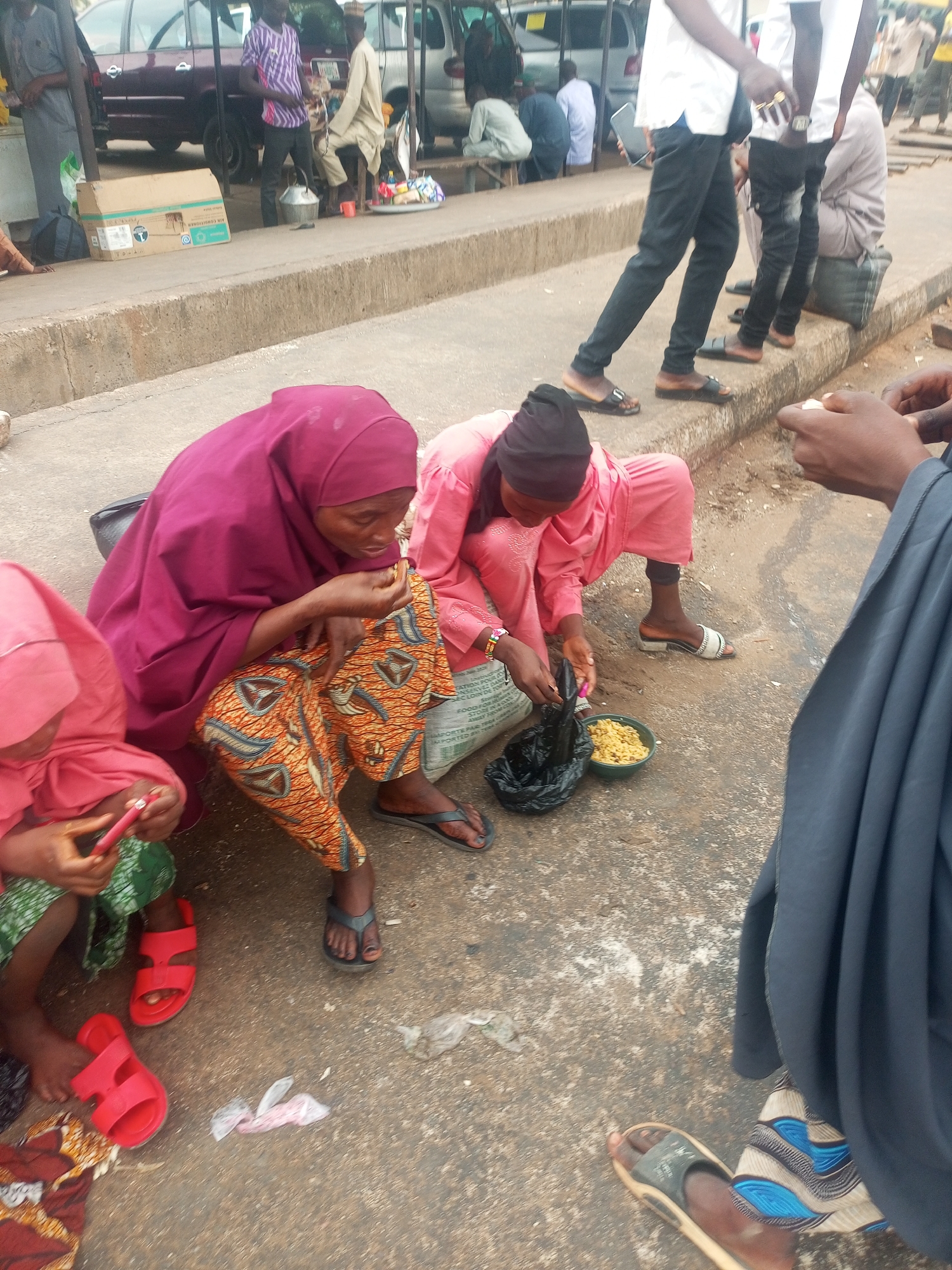 In Gombe, Nigeria, several individuals are situated near a roadside. Two women, dressed in pink and maroon hijab, are seated on the ground. The woman in pink is holding a black plastic bag. Beside them is a child in pink attire, holding a red object. Food is placed in a bowl. Nearby, a group of people is standing, and several vehicles are parked further away. The ground is made of paved asphalt.