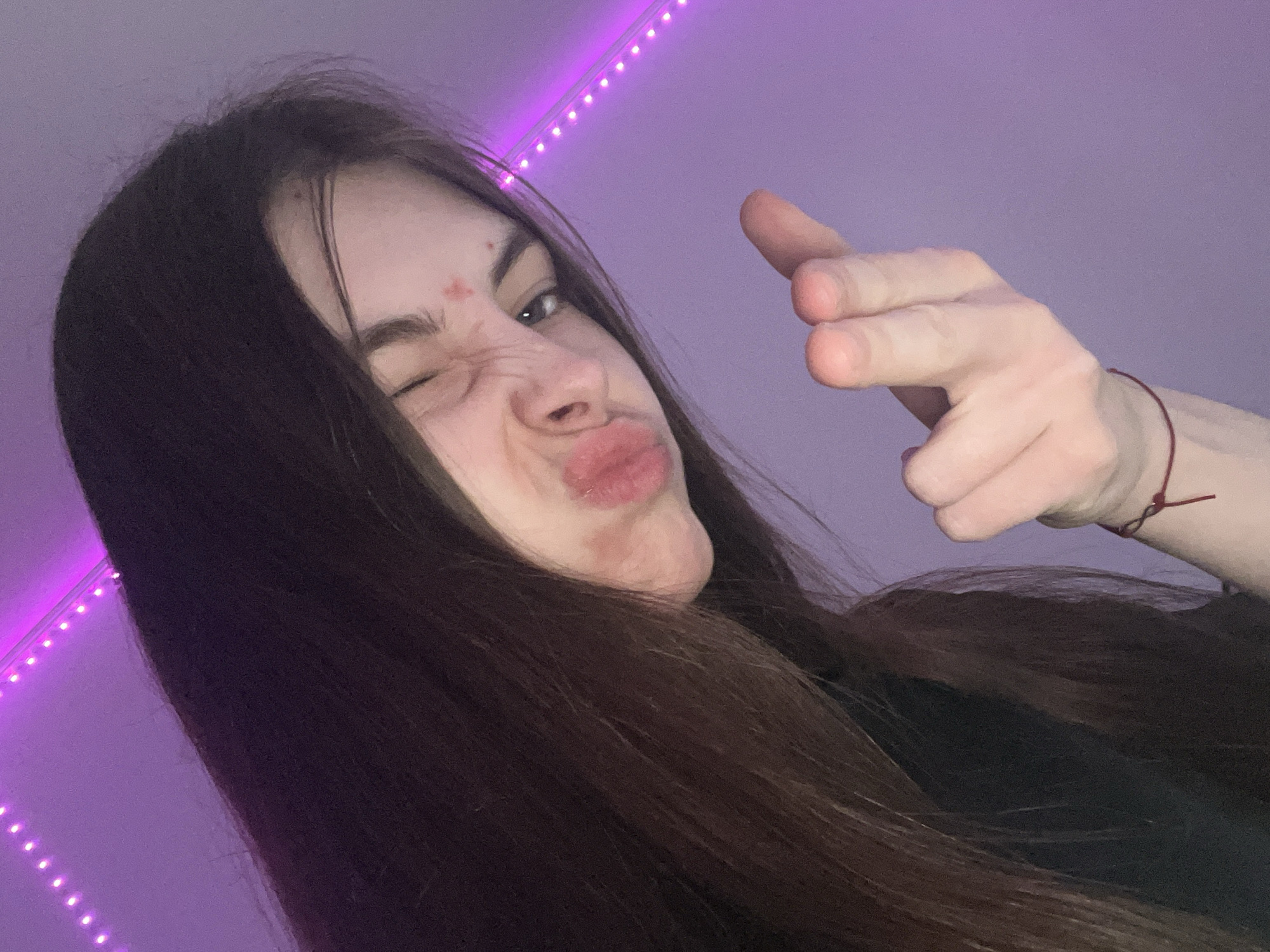 A young person, likely female, is captured in a close-up, casual selfie. She has long, dark brown hair that frames her face and extends out of view. Her expression is playful and exaggerated, with her lips puckered into a "duck face" and one eye squinted or winking, while the other eye is partially visible and looking towards the camera. A couple of small, reddish marks are visible on her forehead and near her eyebrow.
Her right hand is raised and extended towards the viewer, with the index finger pointed forward in a gesture commonly known as a "finger gun." A thin red string bracelet is visible on her right wrist. She appears to be wearing a dark, possibly black, top.
The background consists of a plain light purple wall. Along the top left edge of the frame, a strip of purple LED lights is visible, casting a vibrant glow and serving as the primary light source for the indoor setting. The lighting is artificial and creates an informal atmosphere. There is no visible text in the image. The overall mood is lighthearted and informal.