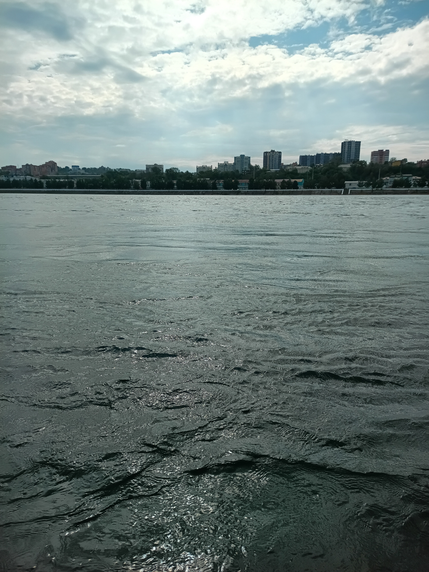 The image shows a wide, choppy body of water in the foreground, likely the Angara River in Irkutsk, Russia. The water is dark and reflects the light, creating a textured surface with many small waves and ripples. In the middle ground, a tree-lined embankment separates the river from a cityscape. The city in the background consists of a mix of residential buildings, some appearing to be apartment blocks, and scattered trees. The sky above is partly cloudy, with patches of blue sky visible between dramatic, streaky clouds. The overall impression is of an urban riverfront scene on a bright but overcast day.