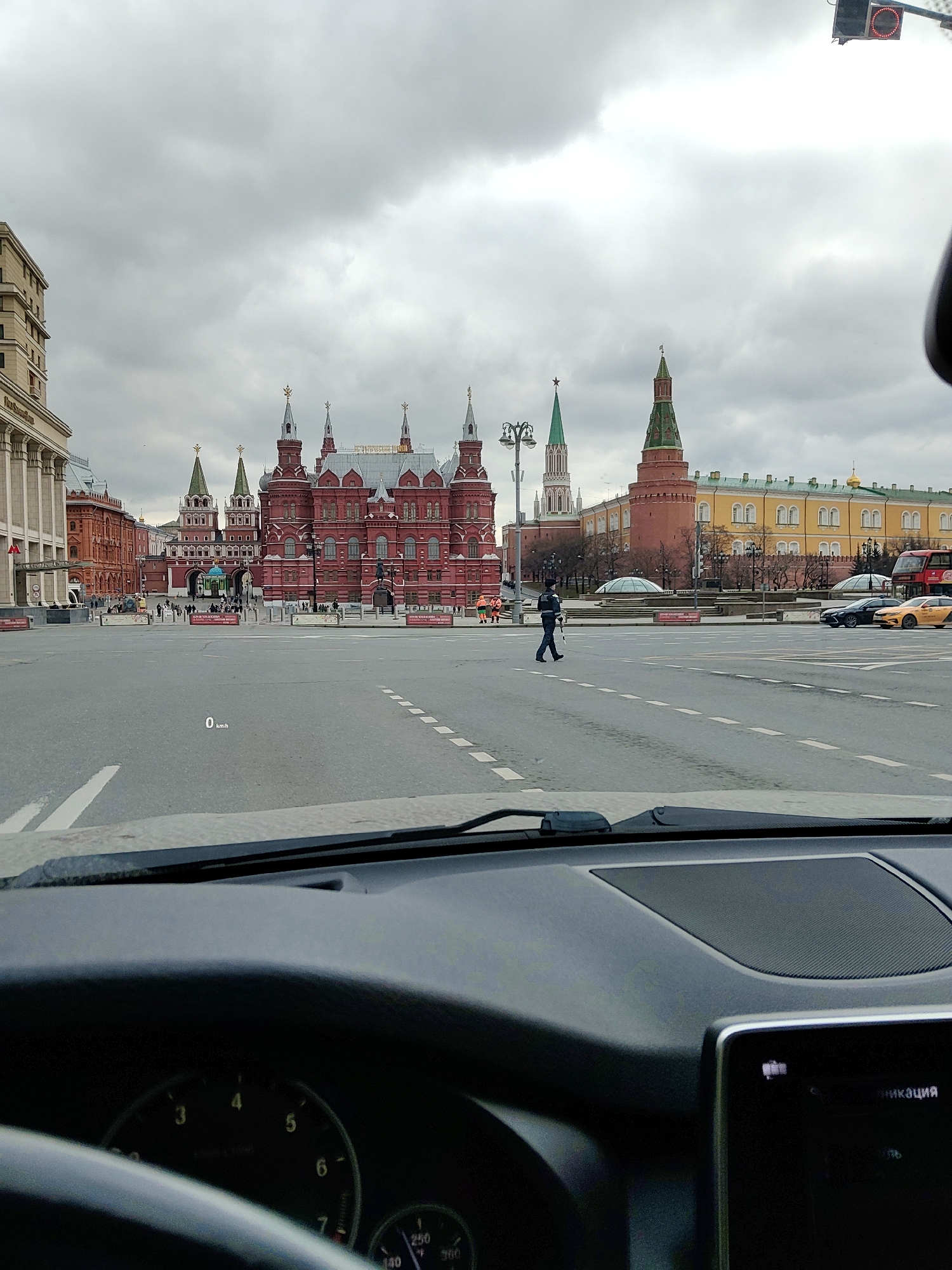 The image is taken from inside a car, showing a view of Red Square in Moscow, Russia. The road in front of the car is mostly empty. In the distance, the State Historical Museum stands out with its red brick facade and multiple towers. To the right of the museum, parts of the Kremlin can be seen, including towers and walls painted in shades of red and yellow. A traffic policeman is walking across the street in the middle of the scene. Several vehicles are also visible on the road to the right. The sky is overcast. The dashboard and steering wheel of the car are in the foreground. The car's speedometer reads "0 kmh", and a display screen is partially visible on the right.