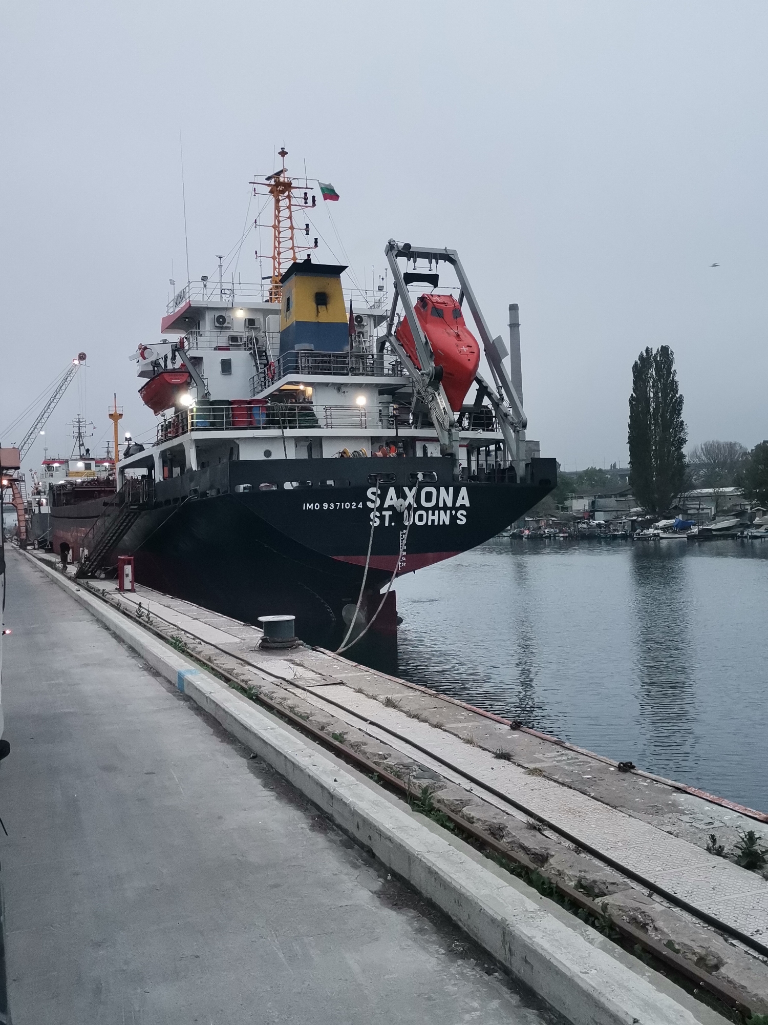 The image depicts a cargo ship named "SAXONA St. John's" docked at a pier in Varna, Bulgaria. The ship, painted black with a red waterline, is securely moored to the pier with thick ropes. Its superstructure features a yellow and blue section near the top, and a Bulgarian flag is visible. Lifeboats are mounted on the ship's deck.
The pier is made of concrete and has railroad tracks embedded in its surface, indicating its historical use for cargo handling. Vegetation grows sporadically along the edges of the pier. The water in the harbor is calm. In the background, across the harbor, there are other boats, buildings, and a tall, slender tree. The sky is overcast, suggesting a cloudy day. The overall environment appears to be a working port area.
