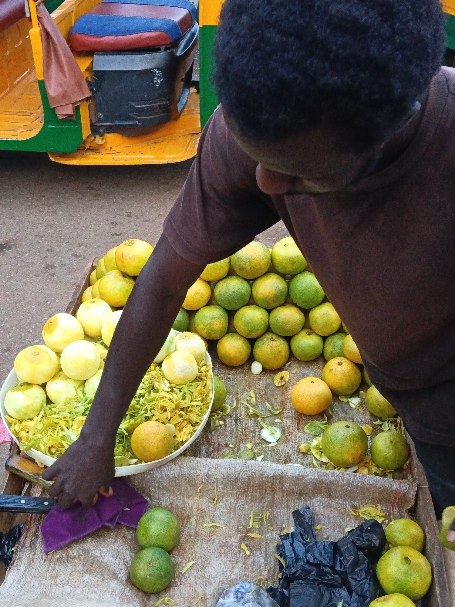 A person, seen from a high angle, is attending to a cart filled with fruits, likely oranges and possibly limes, in an outdoor market. The cart is yellow and green, with a blue and red seat. The person is wearing a brown t-shirt and has dark, curly hair. They are reaching into a large white bowl filled with shredded yellow material, possibly citrus peel, and placing a yellow fruit into it. The cart is laden with piles of whole oranges, some yellow and some green, with some citrus peels scattered on the ground around the cart. A purple cloth and a knife are visible on a burlap surface in the foreground. The overall setting appears to be a street market.