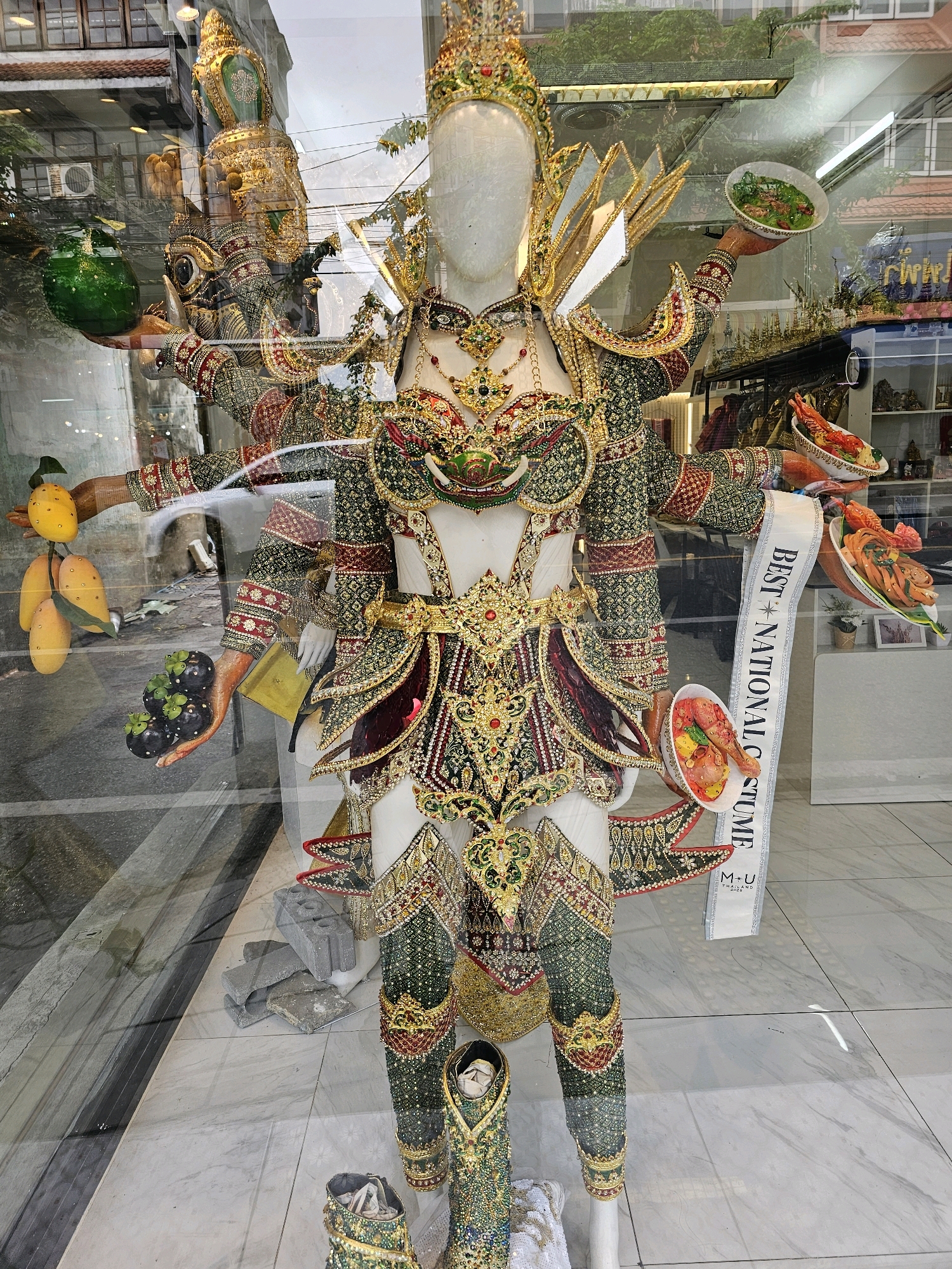 This is a medium shot photograph taken through a glass window, likely in Bangkok, Thailand, as indicated by the surrounding architecture and signage. The main subject is a life-sized mannequin dressed in an elaborate, traditional Thai costume. The costume is richly decorated with gold embroidery, colorful beads, and intricate patterns in green, red, and dark blue. It features a tall, ornate headdress and multiple arms, some of which are holding small dishes of artificial food items like fruits and cooked dishes. A white sash with the text "BEST NATIONAL COSTUME" is draped across the mannequin's torso. The mannequin is positioned in what appears to be a shop window display, with other mannequins and merchandise visible in the background through reflections in the glass. The floor is tiled with a marble pattern.