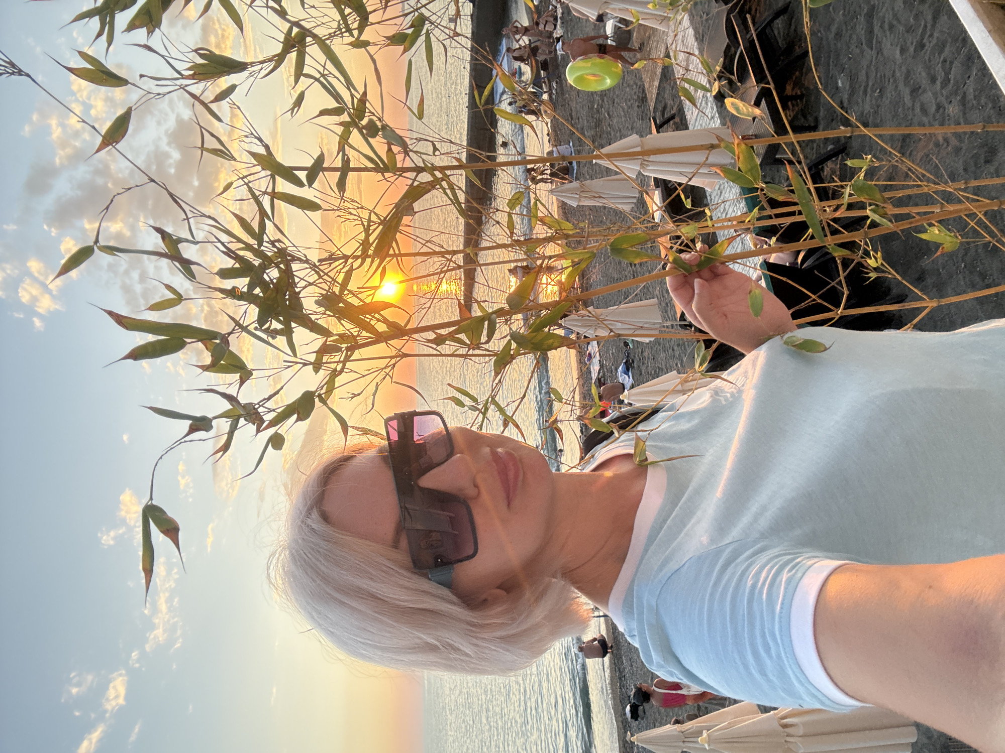 The image depicts a woman with blonde hair and large sunglasses taking a selfie on a beach. She is wearing a light blue t-shirt with a white trim around the collar and sleeves. In the background, the sun is setting over the sea, casting a warm glow. The beach is sandy and lined with rows of closed white umbrellas and dark lounge chairs. Several people are visible on the beach, some sitting or lying down, while others are near the water's edge. The sea appears calm. The location is the city of Sochi, Russia, during sunset.