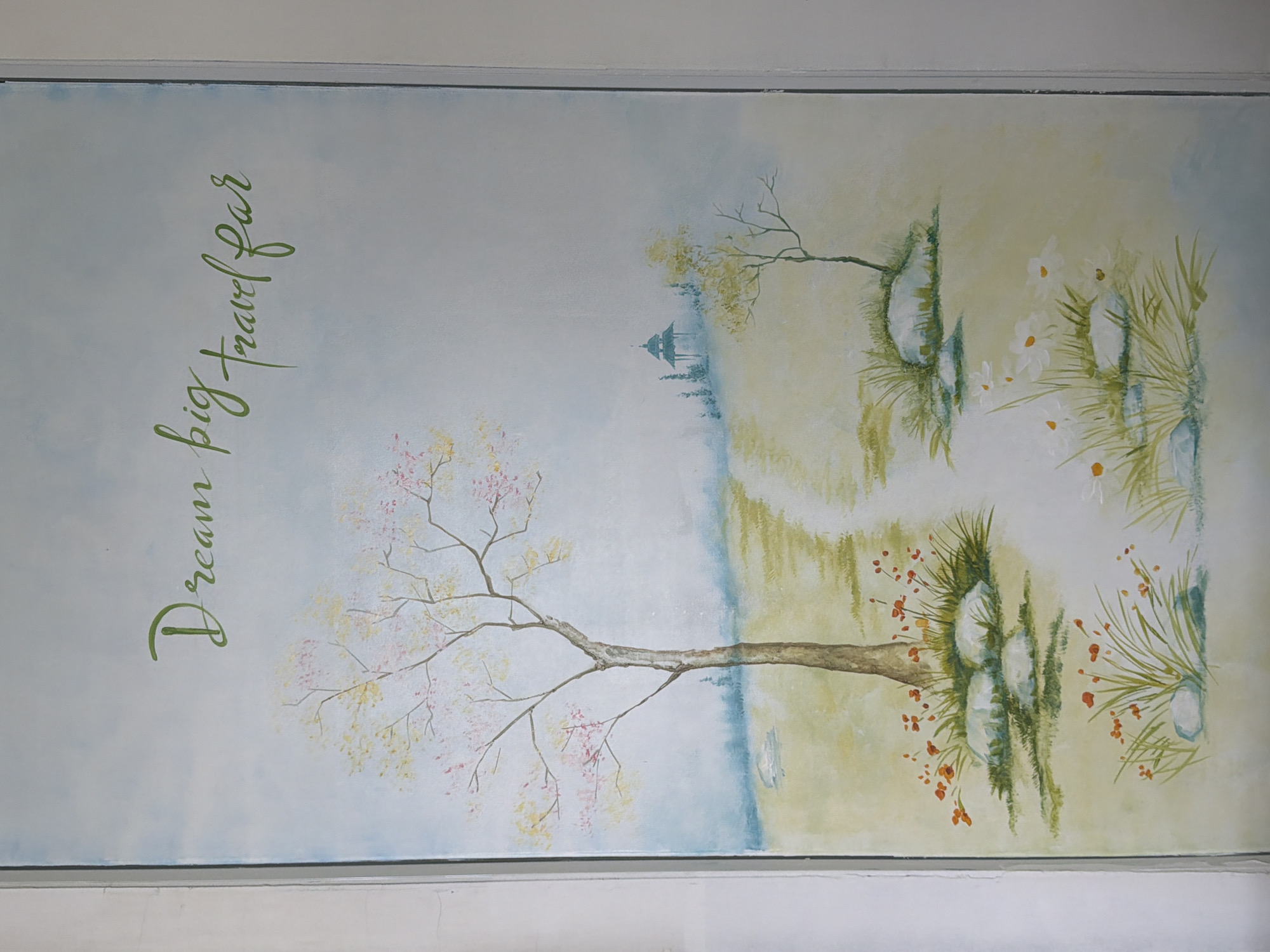 The image is a painting on a wall. The painting depicts a landscape with a path leading through a field. There are trees with pink and yellow blossoms, small orange flowers, white daisies, and rocks scattered throughout the scene. In the background, there is a small pagoda-like structure. Green cursive text that reads "Dream big travel far" is painted at the top of the image. The painting is framed by white borders. The location in Đà Nẵng, Vietnam is not specified by the image.