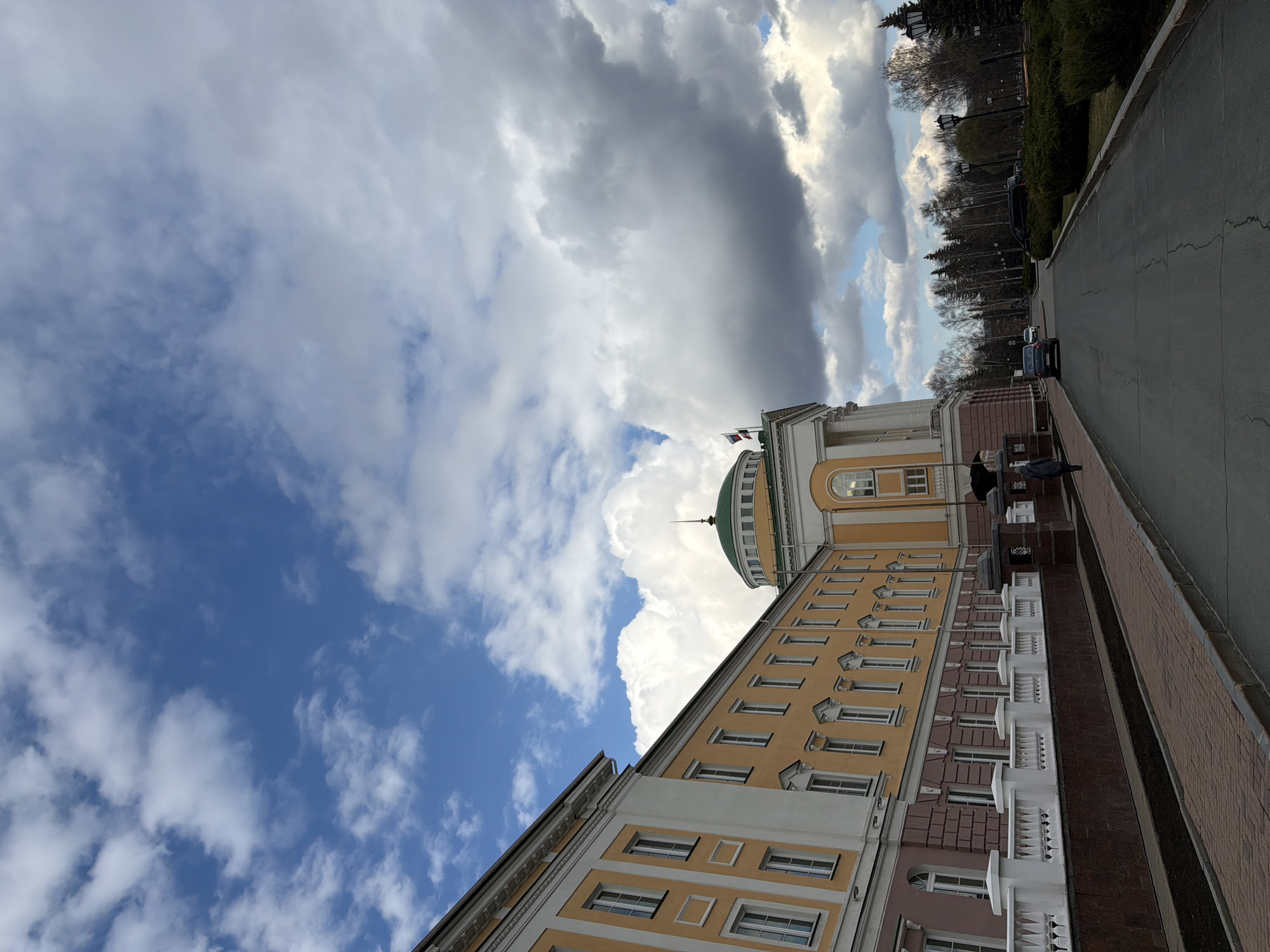 This media file captures a street-level view of a significant building in Izhevsk, Russia, under a dynamic sky. The image is taken during the daytime, likely late morning or afternoon, given the varied light.
The primary subject is a large, multi-story building with a facade predominantly colored yellow and white. The lower sections of the building display reddish-brown and white architectural details, including decorative balustrades along a raised platform. Numerous windows, some with ornate frames, are visible across its face. A prominent green dome topped with a spire rises from the building's roof towards the left, indicating a formal or governmental structure. Near the dome, a flag mast displays what appear to be Russian flags.
In the foreground, a wide, paved area, likely a sidewalk or promenade, runs parallel to the building. This pavement features sections of dark brick-like texture and smoother dark gray asphalt. A few individuals are visible on this pavement; one person dressed in dark clothing stands near a stone balustrade.
Beyond the pavement, a road with visible cracks stretches into the distance, with several cars, including a dark SUV, parked or driving. To the right of the road, a line of trees, some deciduous with bare branches and others evergreen, forms a natural boundary, suggesting an adjacent park or green area. Hedges are also present along the roadside.
The sky is a notable feature, displaying a dramatic mix of bright blue patches and large, contrasting white and dark gray cumulus clouds. The interplay of light and shadow across the clouds suggests a partly cloudy and potentially changing weather pattern. No discernible text is visible within the frame. The overall scene depicts a calm urban environment with impressive architecture.