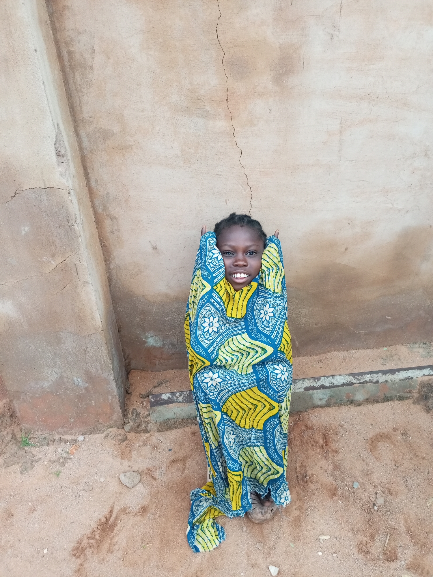 A young girl stands wrapped in a colorful patterned cloth in front of a textured, tan-colored wall. The cloth features a repeating design of blue and yellow abstract shapes and white floral elements. The girl's face is visible at the top of the cloth, and she is smiling with her teeth showing. Her dark hair is braided. Her hands are emerging from the cloth, and she appears to be holding it around herself. The ground beneath her is sandy, with some scattered small stones and a few blades of grass. A section of what appears to be a foundation or low wall is visible at the bottom of the image, running horizontally across the scene. The overall setting suggests an outdoor environment, possibly in a village or rural area, consistent with the informal architecture and ground cover. The image has been taken from a slightly elevated, straight-on perspective.