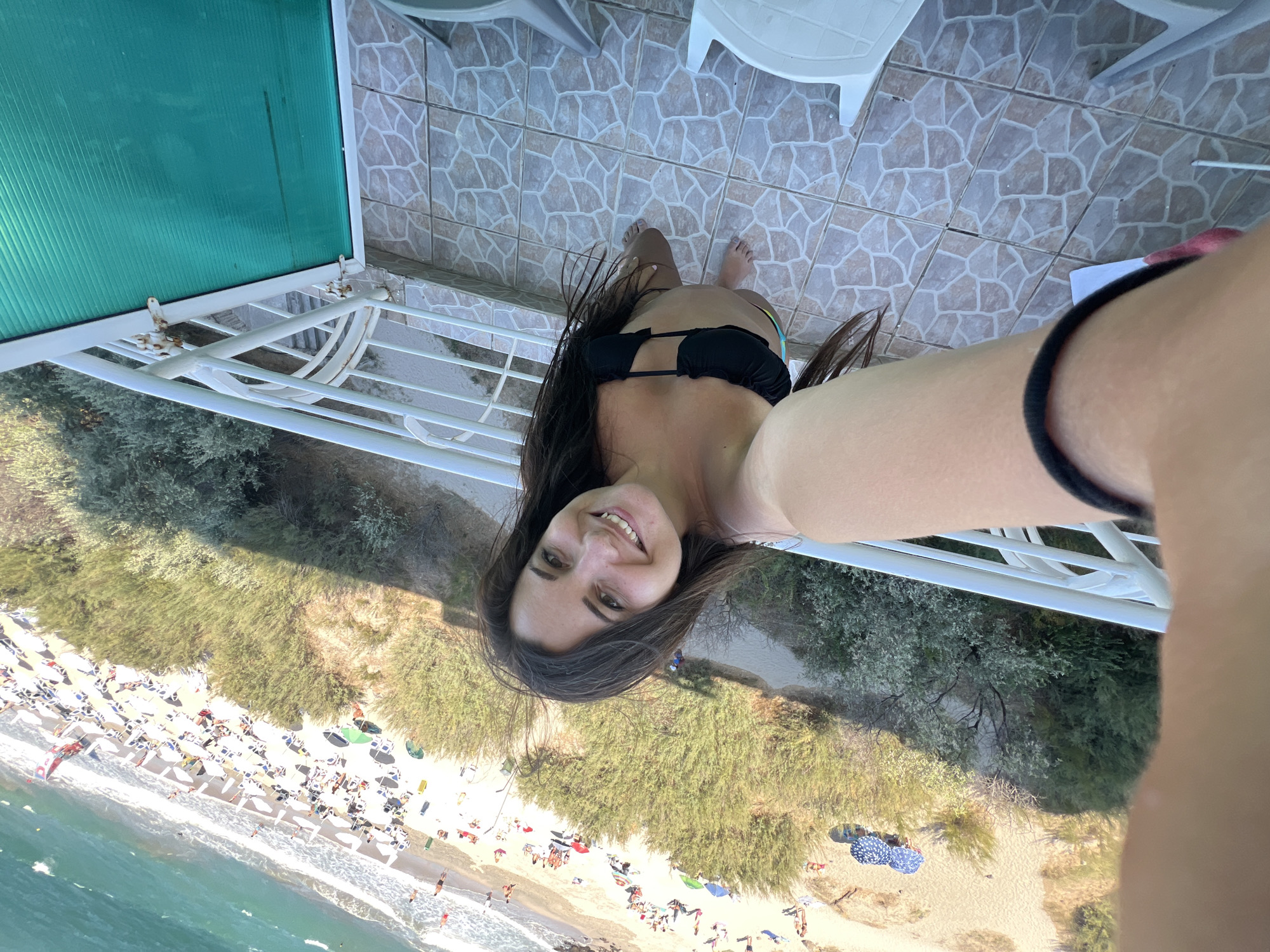 A woman with long, dark hair wearing a black bikini poses for a selfie on a balcony overlooking a beach. The beach is populated with many people lounging on chairs under umbrellas. The sea is visible in the background, with turquoise water and white waves. The balcony has a white railing and a tiled floor. The location appears to be the city of Pomorie, Bulgaria, known for its beaches and natural beauty.