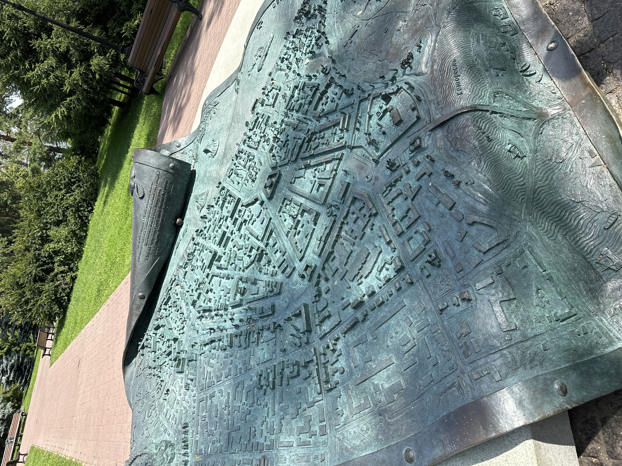An outdoor, eye-level, full shot captures a detailed bronze relief map of a city. The map is set in a park or public space, with a paved walkway and green grass visible in the background. Several wooden park benches are also present. The map itself depicts a dense urban layout with intricate buildings and streets. A notable feature of the map is text inscribed on a rolled scroll element, partially unrolled across the city depiction. The map is weathered, with a patina that gives it a greenish-blue hue. The terrain depicted includes rolling hills and a body of water, suggesting the city's topography. The word "Которосль" is clearly visible on the map, indicating a location, likely a river or district. The scene is illuminated by natural sunlight, casting shadows on the map and surrounding area, suggesting it is daytime.