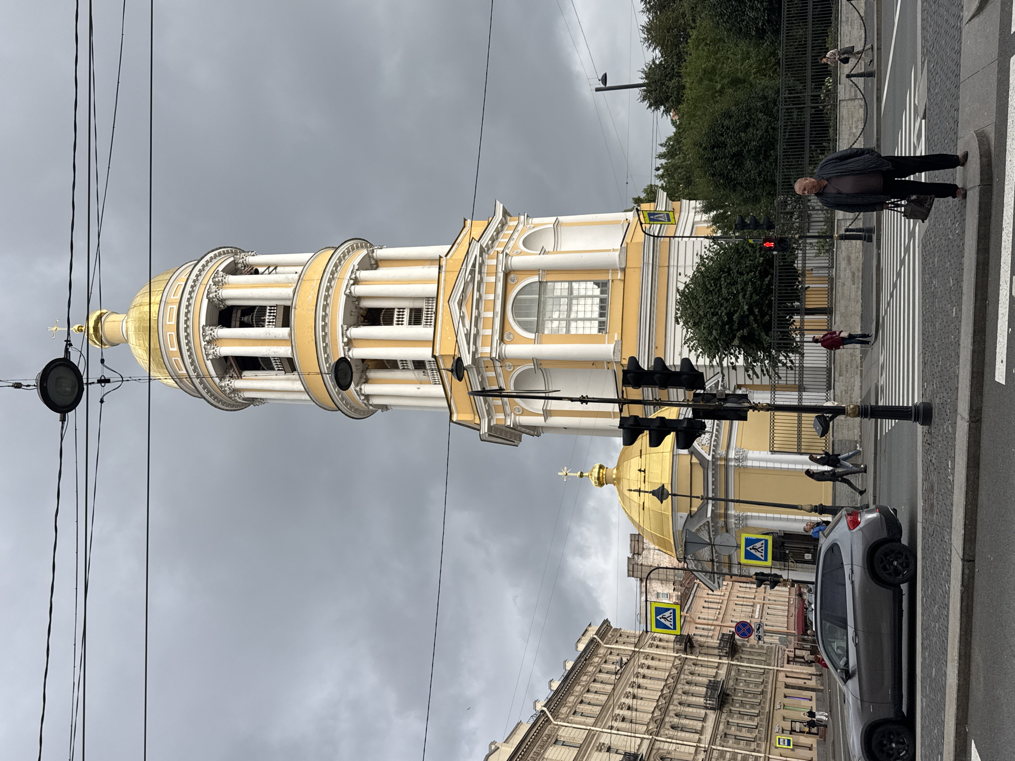 This is an outdoor, eye-level, full-shot photograph of a street scene in St. Petersburg, Russia. The dominant feature is a tall, yellow and white bell tower with a golden dome and cross. In the foreground, a grey SUV is driving past a pedestrian crossing. A man in a dark jacket and trousers is standing on the curb, holding a bag, also near the pedestrian crossing. Several other pedestrians are visible in the midground, some crossing the street, and others walking on the sidewalk. Buildings with classical architectural styles line the street on the left side of the image. The sky is overcast and grey. A traffic light for vehicles shows a red signal, while a pedestrian signal displays a white walking person. The scene appears to be at an intersection with multiple traffic lights and pedestrian crossing signs.