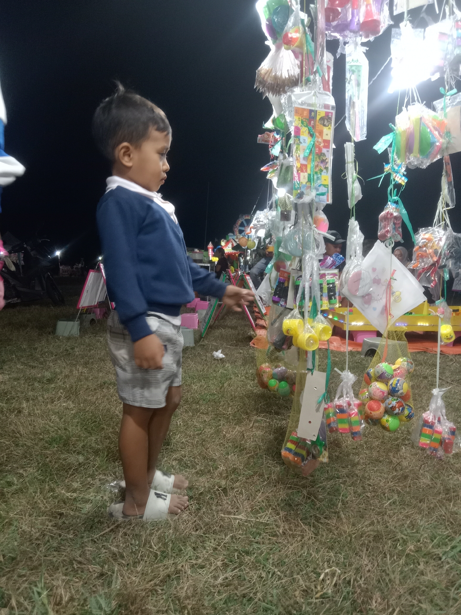 A young boy stands on dry grass, looking at a stall filled with colorful toys and candies. He wears a blue long-sleeved shirt, white collared shirt, and plaid shorts. His white sandals are visible. The stall, illuminated by bright lights, is adorned with various items wrapped in plastic, including small balls, candies, and other trinkets. In the background, other people and stalls can be seen, suggesting a lively night market or fair in Stiaji, Indonesia. The overall scene depicts a child's wonder at a festive outdoor market.