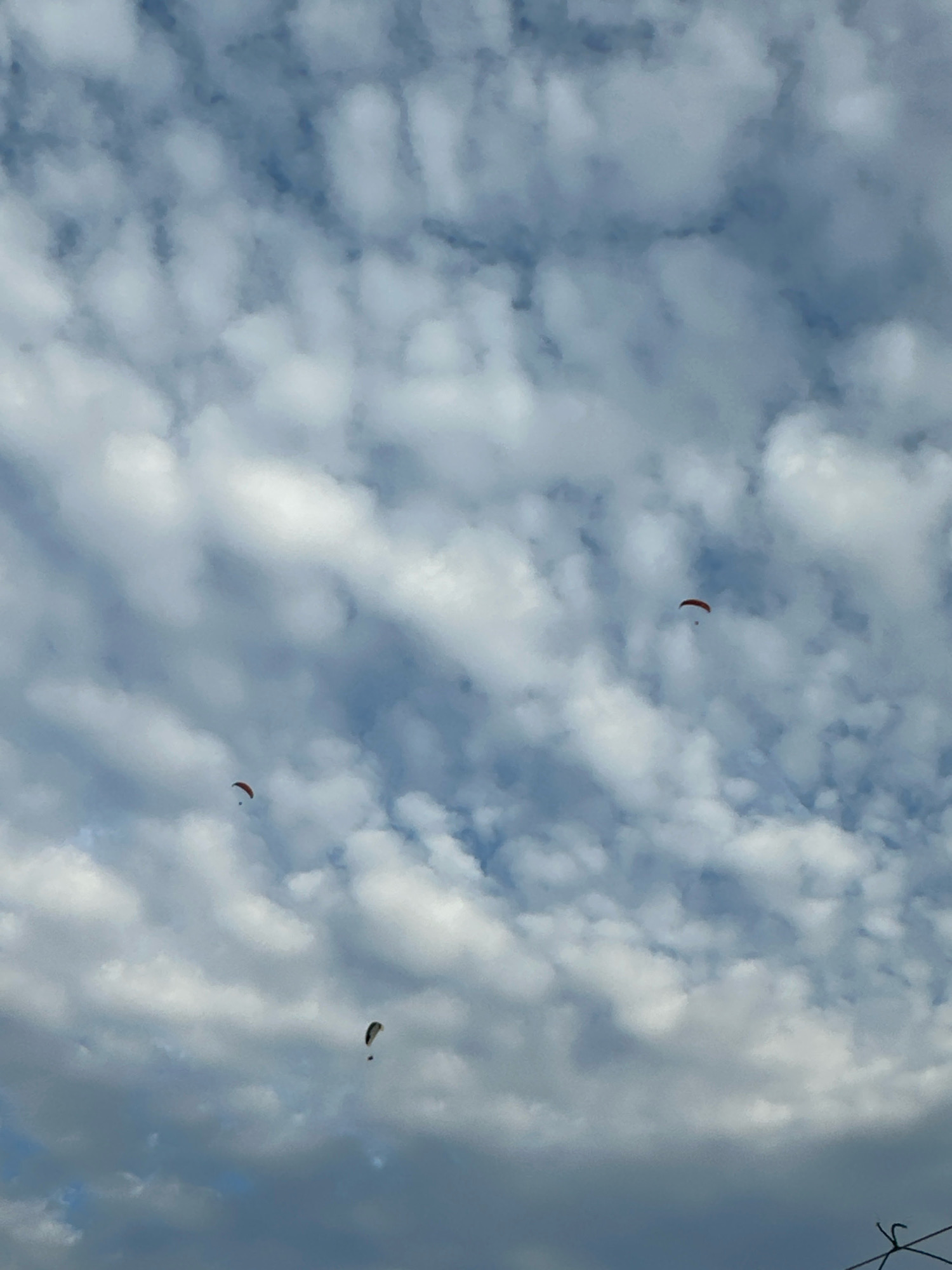 The image captures a cloudy sky with three paragliders visible. The paragliders are scattered across the frame, appearing as small figures against the vast expanse of clouds. The sky is a mix of blue patches and various shades of white and gray clouds, suggesting a partly cloudy day. The paragliders are engaged in the activity of paragliding, suspended in the air. The scene appears to be an outdoor recreational activity. The image does not provide specific location details within Boreti, Montenegro, other than the general environment of a sky with paragliders.