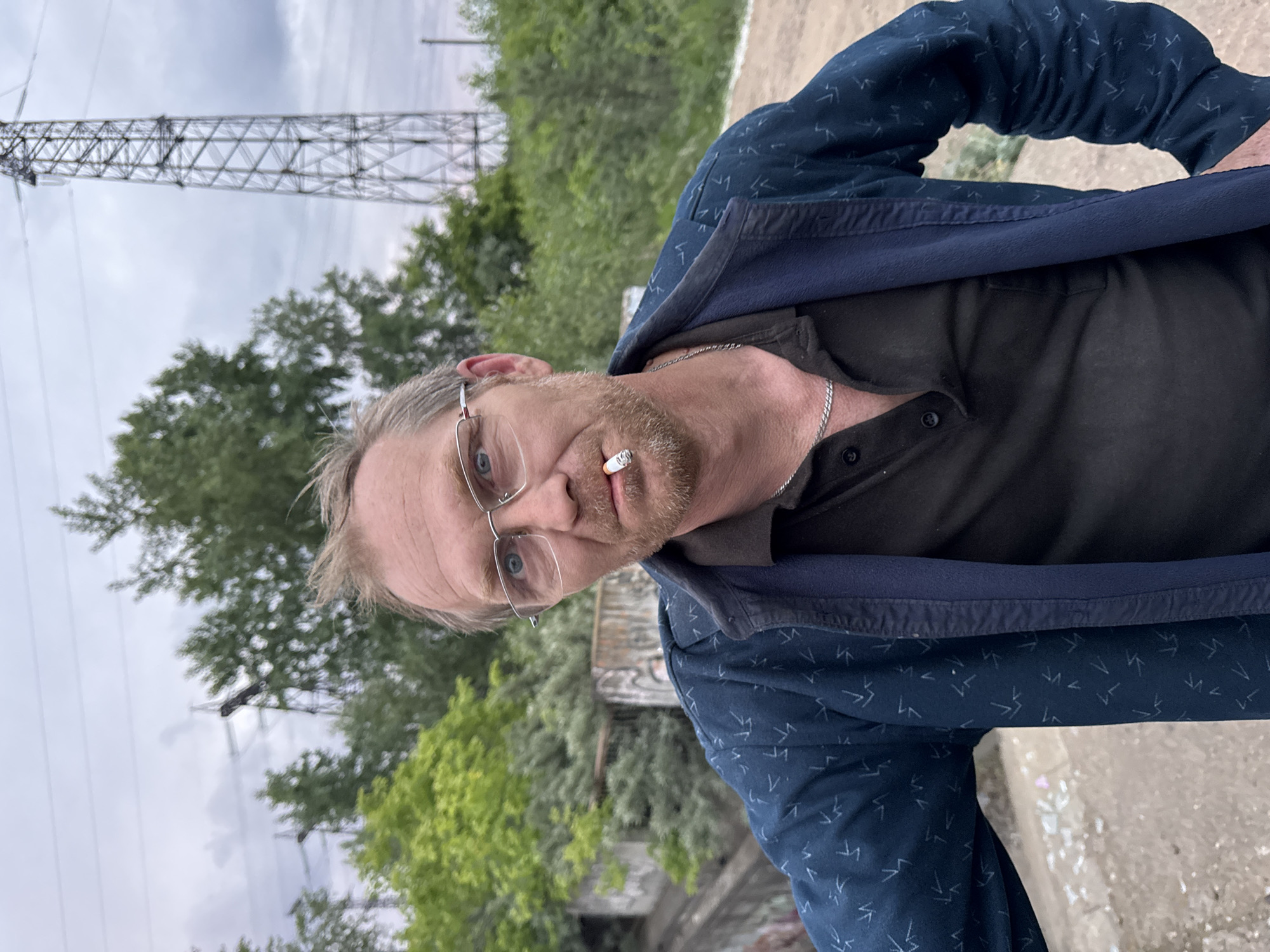 A fair-skinned, middle-aged man with light brown hair and a goatee, wearing glasses and a dark blue jacket over a dark collared shirt, smokes a cigarette. He stands outdoors with a power tower and trees in the background. The location is Chaikovsky, Russia.