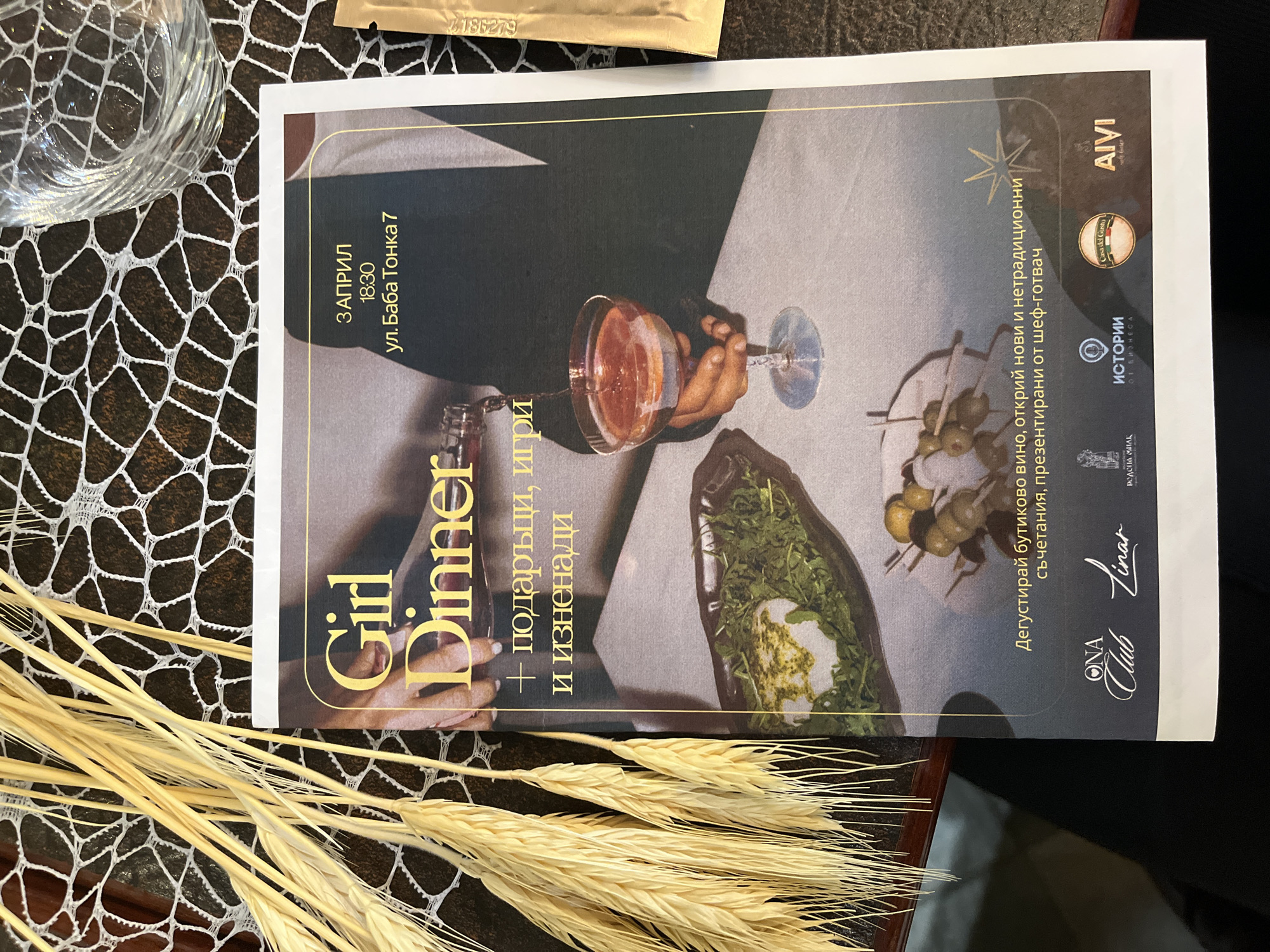 The image shows a promotional flyer for a "Girl Dinner" event to be held on April 3rd at 18:30 on Baba Tonka Street 7 in Varna, Bulgaria. The flyer features an image of a person holding a wine glass, along with food, and includes the phrases "gifts, games and surprises" and "Taste boutique wine, discover new and unconventional combinations, presented by a chef." The flyer also displays logos for ONA Club, Casa del Gusho, Vinar, and AIVI, along with the word "ISTORII" (Histories). The flyer is placed on a dark brown surface near a glass of water and some wheat stalks.
