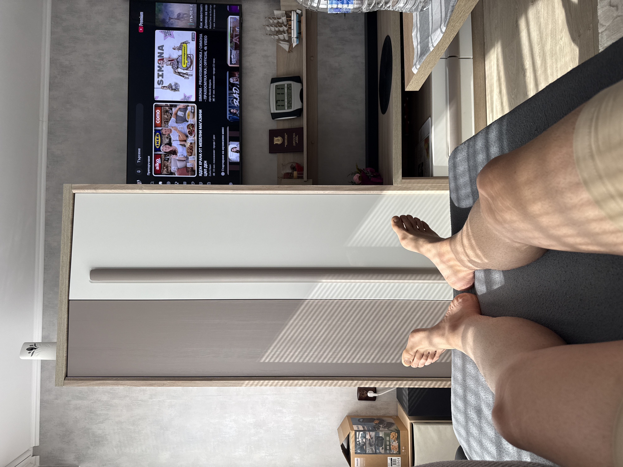 The image shows a person sitting on a gray sofa, with their legs extended. A television displaying content in Bulgarian is visible in the background, along with shelves and a clock indicating the time as 5:24 Sunday, 25th. There is also a diploma, and a cardboard box on the floor. The scene appears to be an interior setting, possibly in Lovech, Bulgaria, given the Bulgarian language on the television. The image captures a moment of relaxation or leisure.