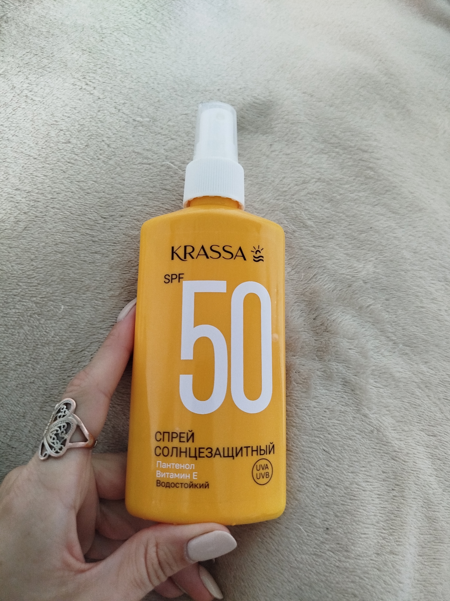 A person's hand, adorned with a silver ring and manicured nails, holds a bright orange spray bottle of sunscreen. The bottle features prominent white text, including "KRASSA" and "SPF 50". Below the "SPF 50", there is Russian text that translates to "sunscreen spray", followed by "Panthenol Vitamin E Waterproof". A small oval shape with "UVA UVB" inside is also visible on the bottle. The background is a textured, light-colored fabric, possibly a blanket or rug. There is no indication of the city of Berdsk, Russia in the image.