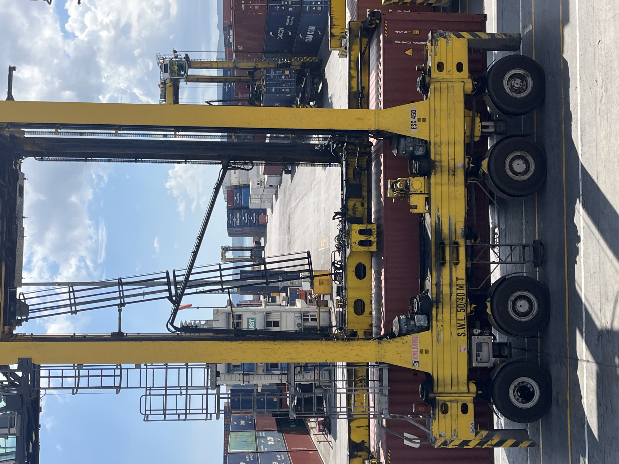 The image shows a port area with a large, yellow straddle carrier in the foreground. It appears to be a bright, sunny day. Numerous shipping containers, stacked in rows, can be seen in the background, indicating a busy cargo handling operation. There are other cargo handling machines visible, some of them appear to be straddle carriers. Text on the equipment reads "KALMAR," "S.W.L. 50/40 MT," "ESC 450," and "RST" The containers are labeled with various companies including "CMA CGM," and "CHA COM." The environment suggests a commercial, industrial setting typical of a port facility.