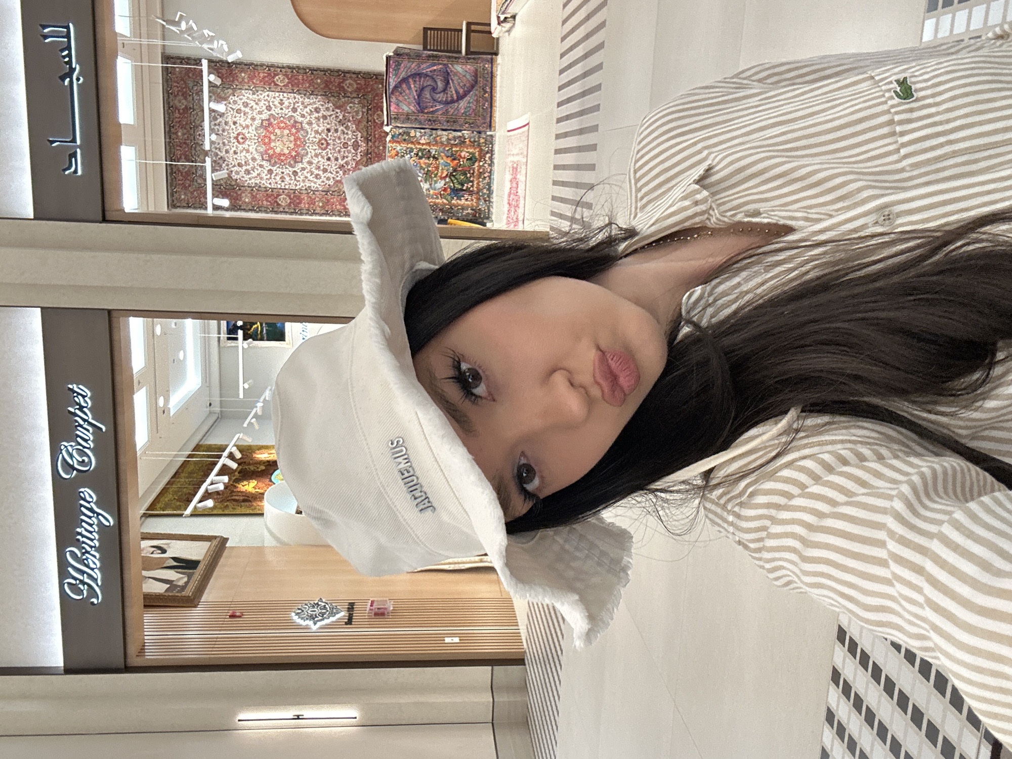 A vertically oriented medium shot depicts a young woman facing slightly right, with her head tilted leftward. She is wearing a white Jacquemus bucket hat and a tan-and-white striped collared shirt. Her dark hair falls over her shirt.
The background shows the interior of a store named "Heritage Carpet", as visible on a sign to the left. Carpets are on display in the store window, and a framed piece of art is visible below. Striped panels are located to the right.
Based on the store's branding and the overall aesthetic, it is likely that the location is in Dubai, United Arab Emirates, known for its luxury shopping and carpet stores. The image suggests an upscale retail environment.