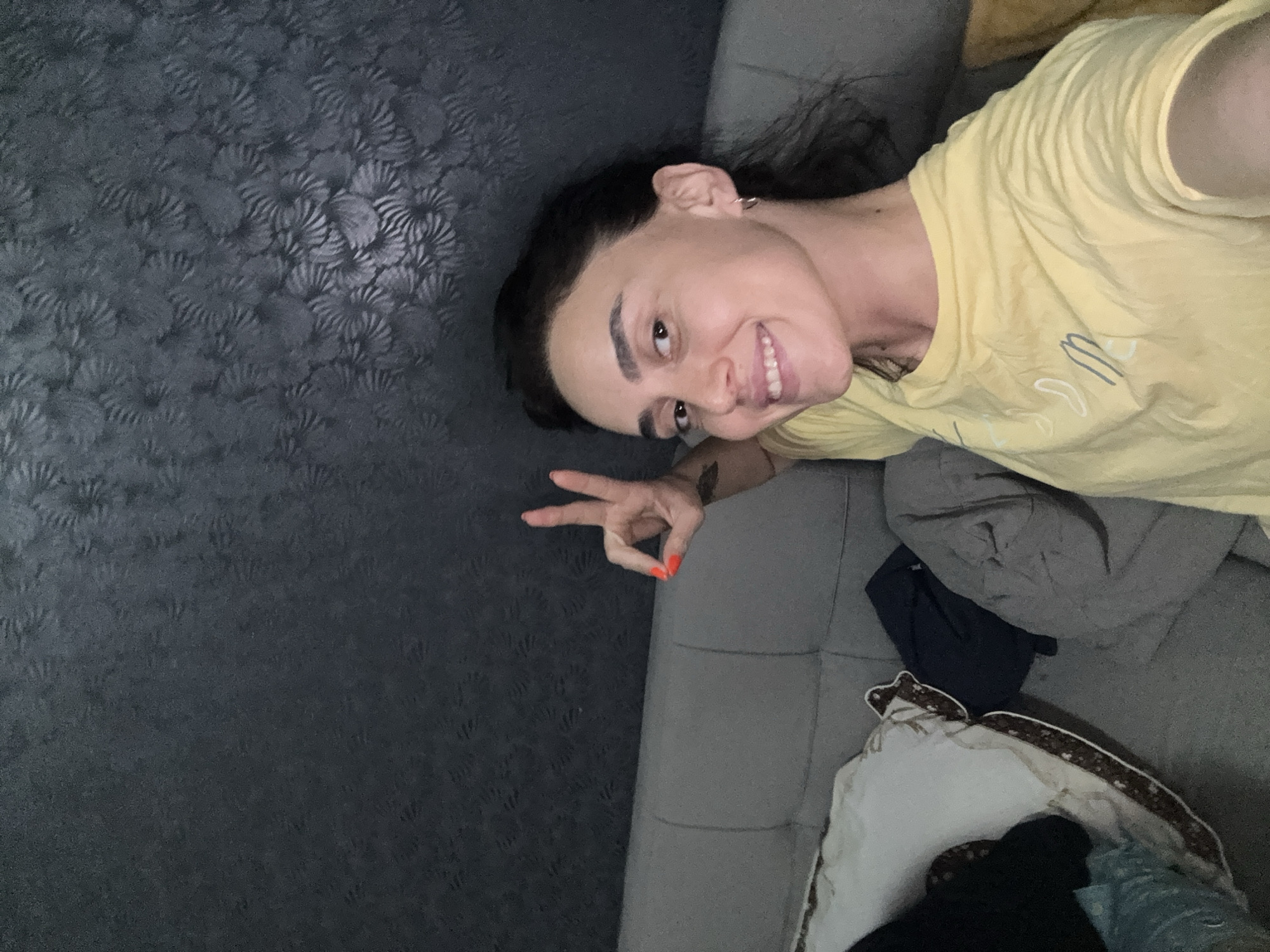 A young woman with dark hair and fair skin is taking a selfie. She is wearing a yellow t-shirt and is seated on a grey sofa. Her right hand is raised, forming a peace sign with her index and middle fingers extended. Her fingernails are painted bright orange. She has a hoop earring in her left ear and a tattoo on her left arm. The background appears to be a textured, dark grey wall. The sofa also has pillows and what appears to be folded clothing on it. The scene is likely indoors, possibly in a residential setting within Tbilisi, Georgia.