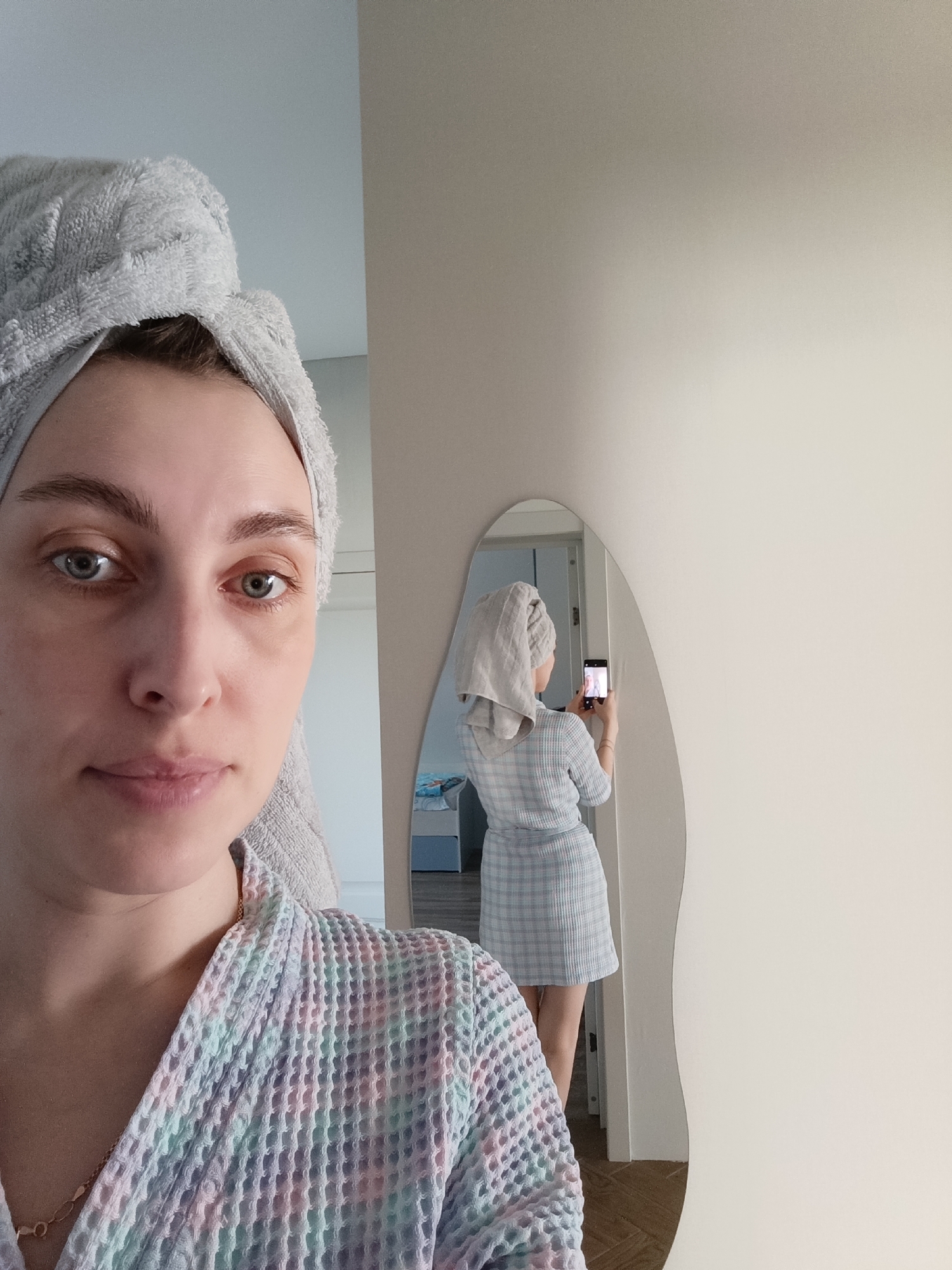 An indoor scene in Chaikovsky, Russia, depicts a young adult female captured through a direct frontal view and a mirror reflection. In the foreground, the woman is seen from the chest up, wearing a pastel multi-colored waffle-knit bathrobe and a light grey terrycloth towel wrapped around her head. She has fair skin, light-colored eyes, and a neutral expression while looking directly at the camera. To her right, an irregularly shaped mirror reflects her full figure from behind. In the reflection, she wears a light blue and white checkered short dress or bathrobe, with the same grey towel on her head. She holds a black smartphone with both hands, positioned to photograph her reflection, with the phone screen displaying her image. The room features light-colored walls and, visible in the reflection, a light brown herringbone patterned floor and an open doorway leading to another room with light walls and a light blue or grey furniture item. The lighting is bright and even throughout the scene.