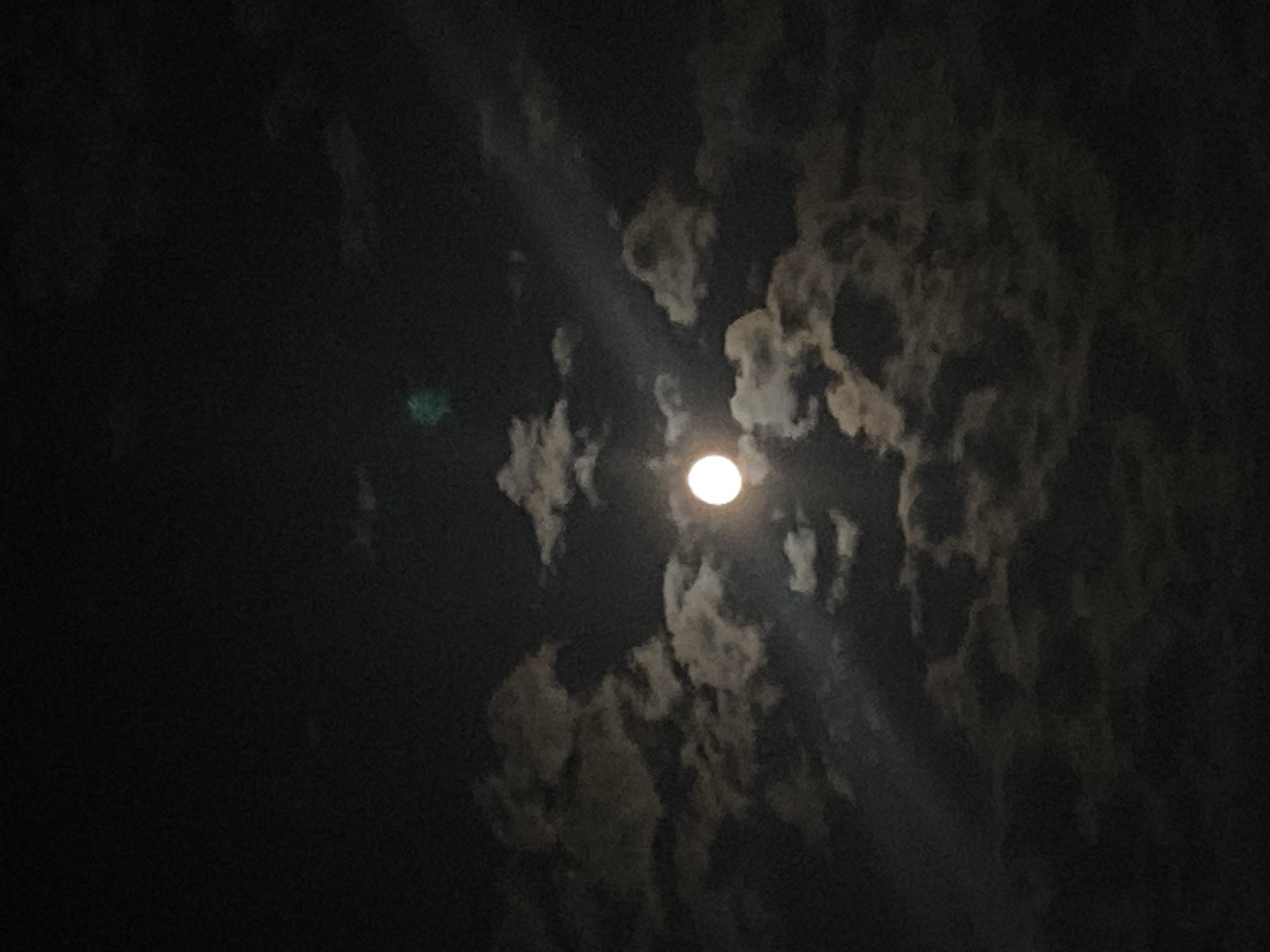 The image is a nighttime photograph of the moon partially obscured by clouds. The moon is a bright, circular object in the center of the frame, emitting a soft glow. Wispy clouds surround the moon, with some appearing darker and denser, while others are lighter and more translucent, allowing the moonlight to filter through. A diagonal beam of light emanates from the moon, cutting across the clouds. In the upper part of the frame, a faint, greenish luminous haze is visible. The overall scene depicts a cloudy night sky with a prominent moon. There are no discernible elements that would allow for the identification of a specific location within the city of Iași, Romania.