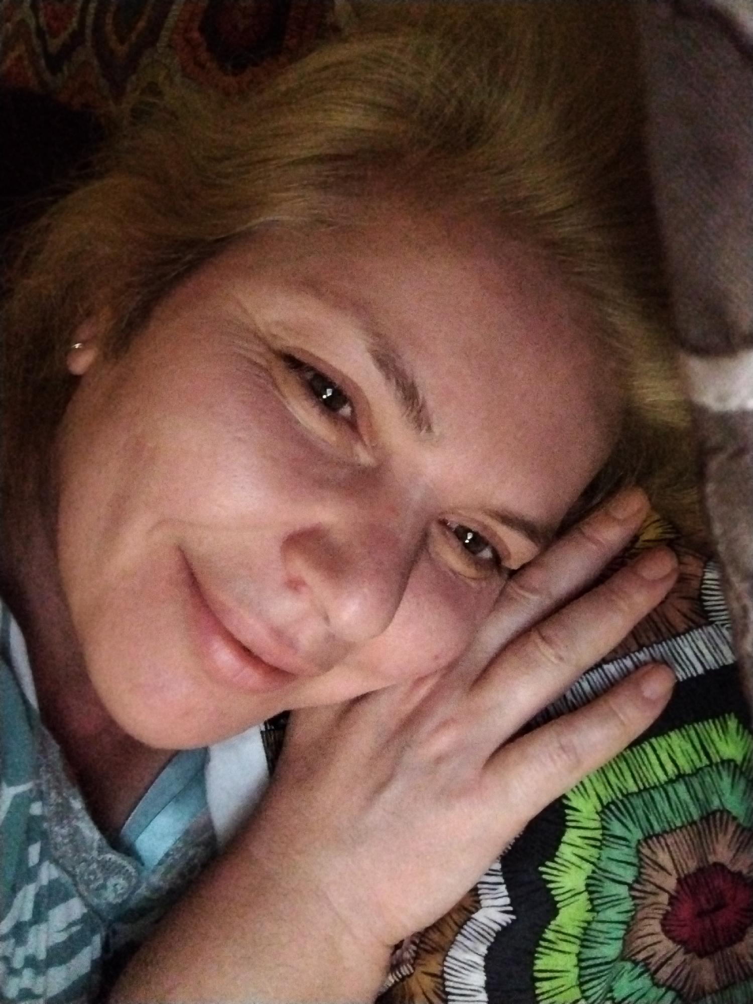 A close-up shot depicts a woman with light blonde hair, lightly wrinkled skin, and brown eyes. She is smiling gently and resting her head on her hand, which is placed on a patterned surface. The surface is a multicolored pillow with concentric circular patterns featuring green, brown, and white designs over a black background. A portion of her shirt, visible on the left, is blue with a grey leafy design. An earring is visible in her left ear. The background is blurred and out of focus. There is no indication of the location being Al Mas'oudi or the United Arab Emirates.