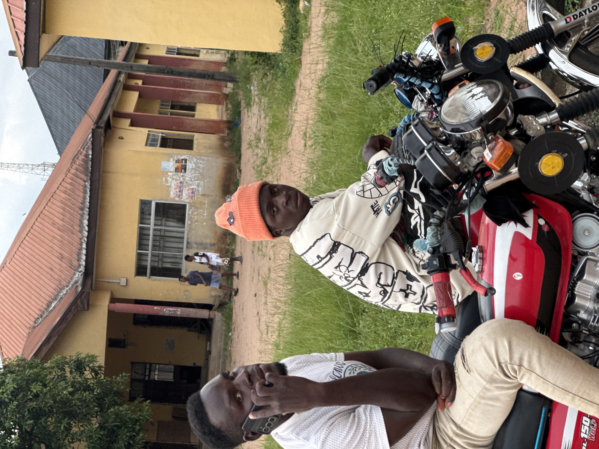 This outdoor image, likely taken in Ihiagwa, Nigeria, features two men on a red and black motorcycle in the foreground, set against a backdrop of buildings and other people.
In the immediate foreground, a man wears an orange beanie and a light-colored jacket adorned with black graphics, including the word "HATE." He looks directly at the camera with a neutral expression. Seated behind him on the motorcycle is another man, dressed in a white textured t-shirt (with faint text like "CHILL") and tan ripped pants. He is actively engaged in a phone call using a black smartphone. The motorcycle, a prominent object in the scene, has "DAYLONG" visible on its front wheel and "DL 150 WOLF" on its side, indicating its model.
The background consists of a dirt path and green grass. Several yellow-painted buildings with dark, corrugated metal roofs are visible, featuring windows and some unreadable posters or flyers on their walls. A sturdy wooden pole or beam stands vertically near the buildings. A tree with lush green foliage is situated in the mid-ground. Further back, two additional individuals can be seen; one appears to be wearing a white and black jersey with the number 23 or 25, while the other is dressed in a dark shirt and light shorts. A faint metal structure, possibly a telecommunication mast, is visible against the sky in the far distance.
The scene is well-lit by natural daylight, suggesting it is daytime, possibly under an overcast sky. The setting evokes a sense of a local community or a semi-urban area.