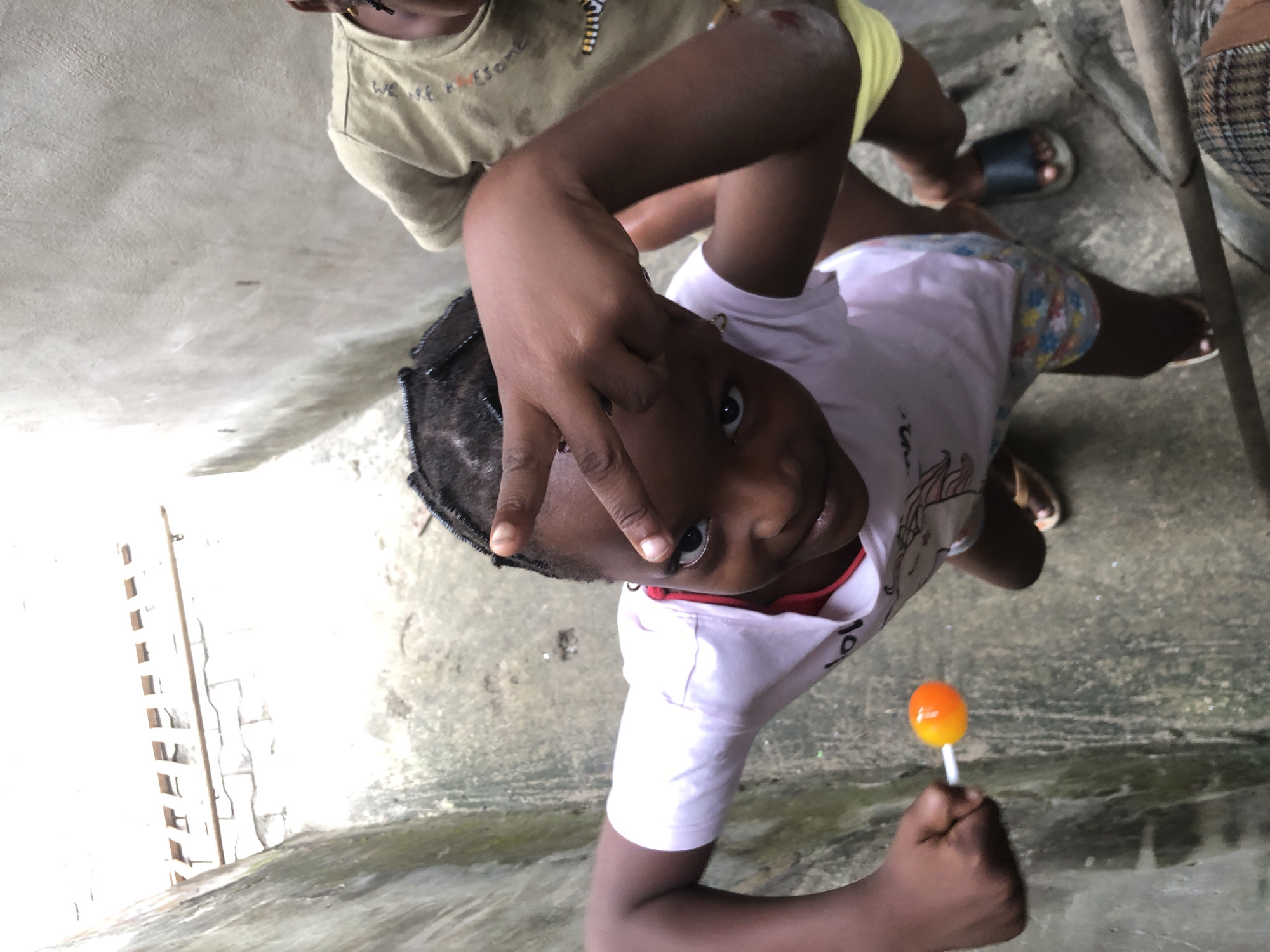 In Ado, Nigeria, two young girls are captured in a high-angle outdoor shot. The girl in the foreground, wearing a pink t-shirt and floral shorts, holds an orange lollipop in her left hand and playfully makes a peace sign with her right hand near her face. Her eyes are directed towards the camera. Behind her, another child in a tan shirt and yellow shorts is partially visible. The environment appears to be a narrow alleyway with concrete walls and a textured ground, suggesting an urban setting. The lighting is bright, casting subtle shadows, and the overall atmosphere suggests a candid moment of childhood play.
