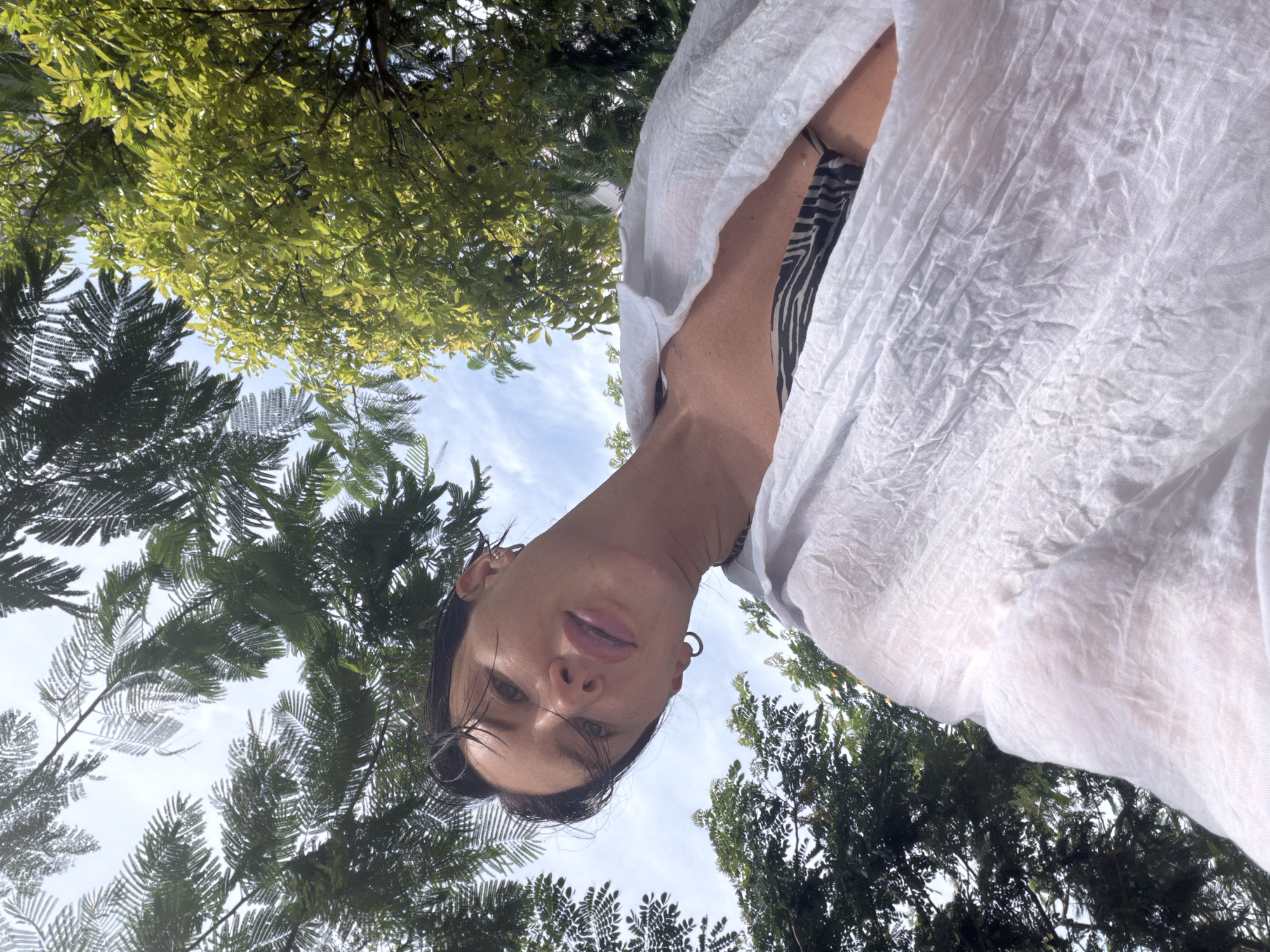 A low-angle, medium shot captures a woman from the chest up, against a backdrop of lush green trees and a blue sky. She is wearing a white, unbuttoned shirt over a black and white patterned bikini top. Her dark hair is slicked back, and she wears hoop earrings in both ears. The woman's face is turned towards the camera, with her lips slightly parted. The image appears to have been taken outdoors, possibly in a tropical location. The specific location in Dong Ninh Hoa Ward, Vietnam is not discernible from the image itself.