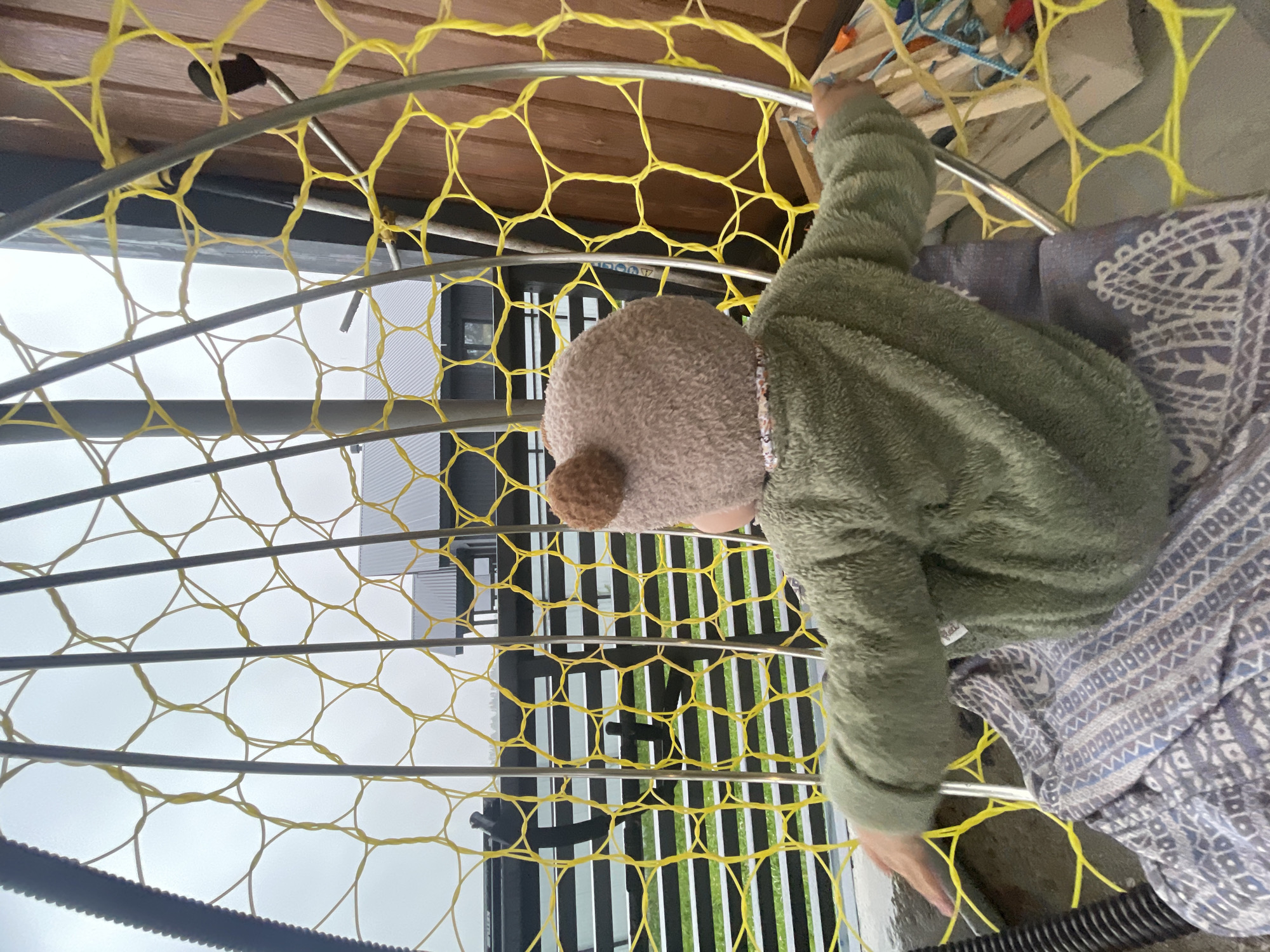 A child wearing a fuzzy green jacket and a bear-eared hat sits in a yellow netted hammock chair on a patio in the city of Podshivalovo, Russia. The child is facing away from the camera, looking towards a black fence and a building with a dark roof. The hammock is made of thick yellow netting and silver metal poles, with a patterned blanket draped over the seat. The patio appears to be made of concrete, and the background suggests an overcast day.