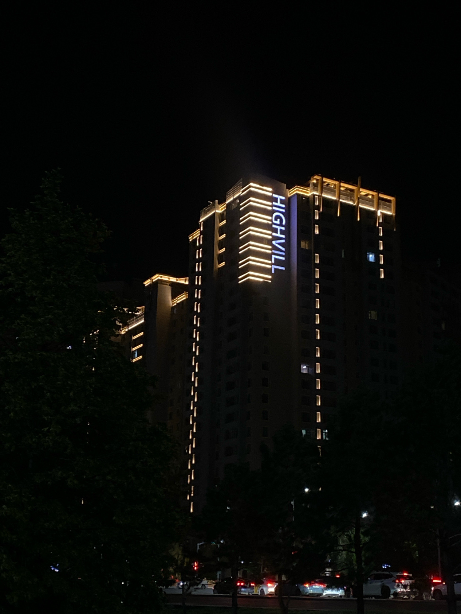The image depicts a nighttime street view in Astana, Kazakhstan, featuring a modern high-rise building with illuminated signage reading "HIGHVILL" in blue. The building is adorned with horizontal strips of warm yellow light accentuating its architectural features. In the foreground, dark silhouettes of trees partially obscure the view of the building and the street. The street below is visible with several vehicles, some with their headlights and taillights illuminated, suggesting traffic flow. The overall scene captures an urban night environment with prominent architectural lighting.