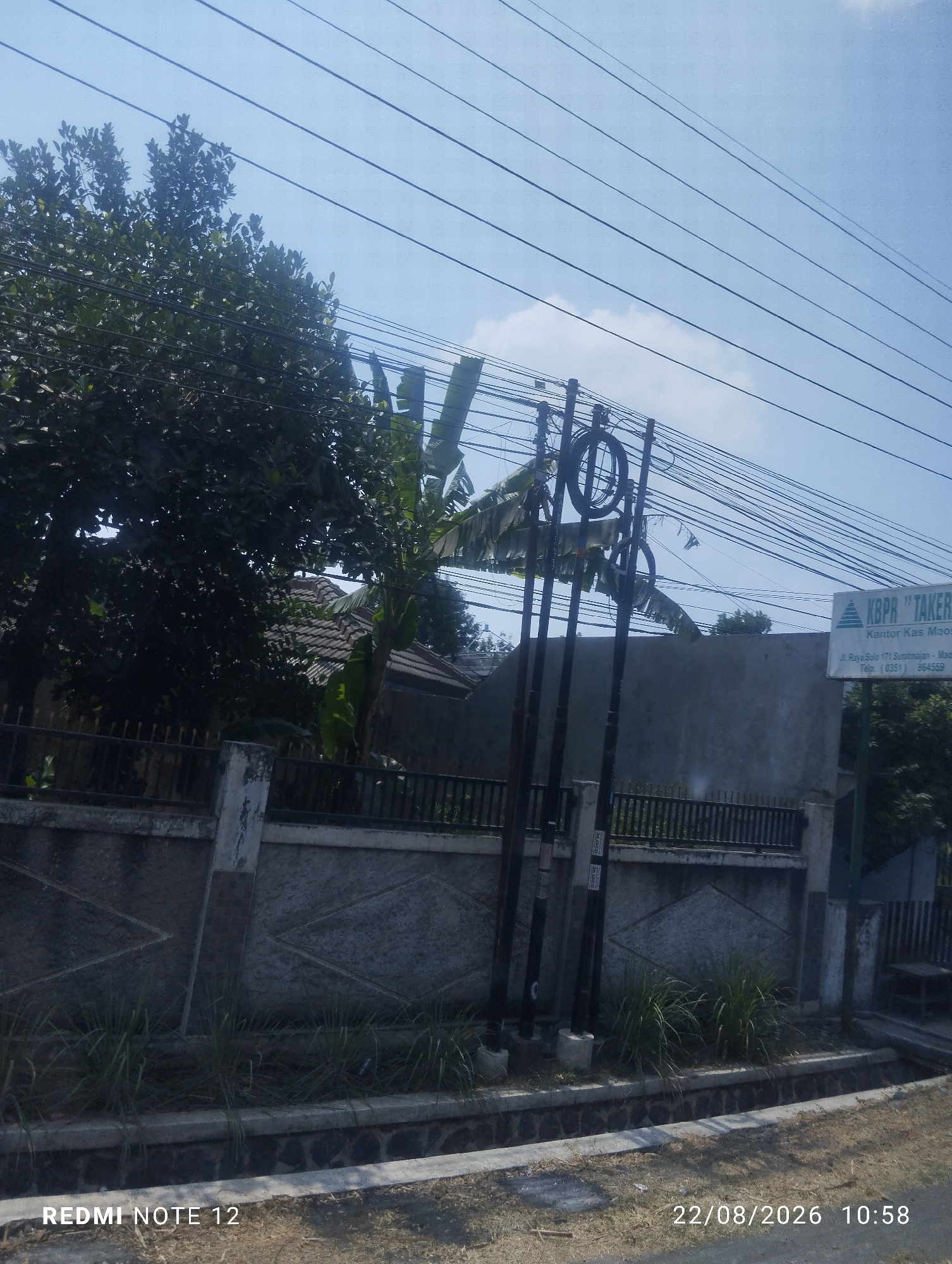 The image captures a street scene in Klagen Gambiran, Indonesia, on August 22, 2026, at 10:58 AM. The foreground features a concrete ditch bordering a dry, grassy area. Beyond this, a textured concrete wall with diamond-shaped patterns serves as a boundary. A black metal fence runs along the top of this wall. Lush green trees and banana plants are visible behind the fence, with a tiled roof of a house peeking through the foliage. Overhead, a dense network of electrical wires crisscrosses the bright blue sky, supported by utility poles. A sign, partially obscured by trees and wires, reads "KBPR 'TAKER'" and displays an address and phone number. The overall impression is of a typical residential street scene in a tropical climate, with evidence of electrical infrastructure and vegetation.