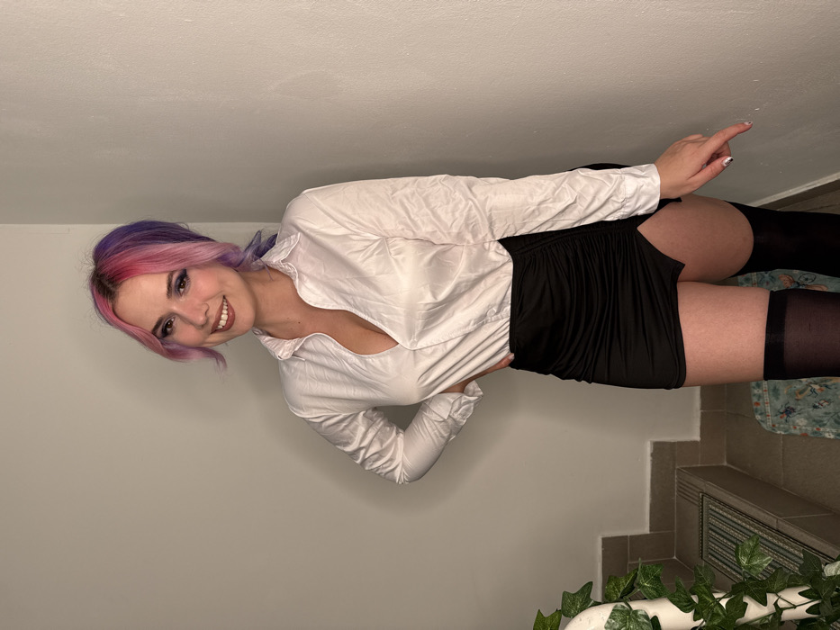 A female subject is depicted standing in an indoor setting, identified as the City of Zagreb, Croatia. She has split-dyed hair, pink on the frontal and top sections, and purple on the rear and underneath, styled with bangs. Her facial expression is a smile directed towards the camera. She is wearing a white long-sleeved collared shirt, unbuttoned to reveal cleavage, a black ruched mini-skirt with a diagonal slit on the right thigh, and black thigh-high stockings. Her right hand rests on her hip, while her left hand is extended downwards, with the index finger pointing at the adjacent wall. The background consists of two plain, light-colored walls forming an interior corner. To the subject's left, a tiled staircase with a white banister, partially covered by green foliage (likely ivy), is visible, along with a patterned mat on a lower step. The scene is illuminated by even, diffused lighting.
