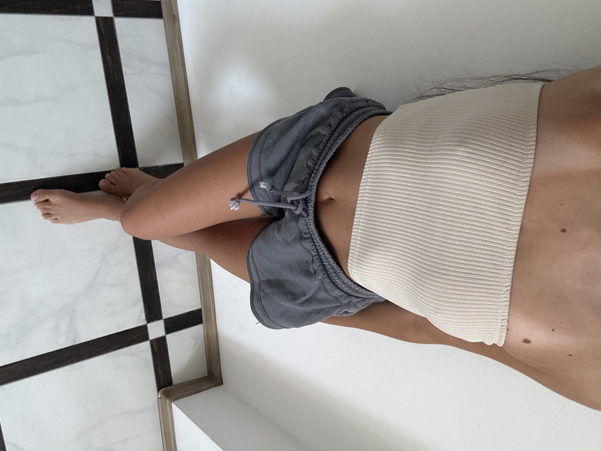 A young woman, viewed from a high-angle, stands leaning against a white wall. She is wearing a beige ribbed bandeau top and grey shorts. Her legs are crossed at the knees and her bare feet are visible on a checkered floor. The floor is made of white marble tiles with dark brown rectangular inlays, forming a grid pattern. A thin wooden trim separates the floor from the wall. The image appears to be taken indoors, possibly in an apartment. There are no visible signs or landmarks that would specifically place this scene in Chișinău, Moldova.