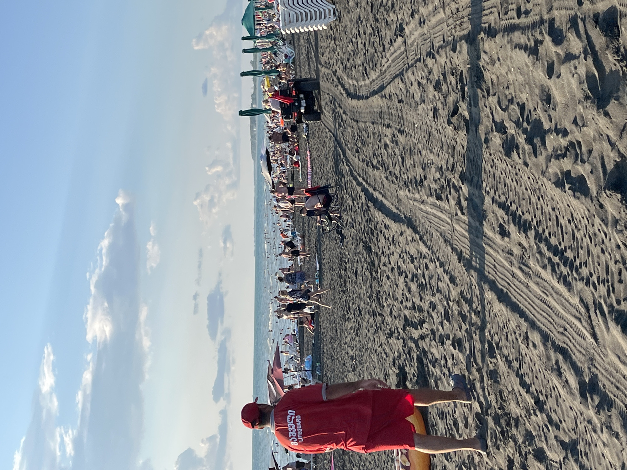 In Ureki, Georgia, a busy black sand beach is depicted on a sunny day. The foreground is dominated by the textured sand, marked with tire tracks from a vehicle, likely an ATV. A lifeguard, clad in a red shirt with the word "LIFEGUARD" and Georgian script printed on it, and red shorts, stands facing away from the camera, looking towards the sea. He is wearing a red cap and blue sandals. To his left, a yellow floatation device is partially visible.
Further back, the beach is populated with numerous people enjoying the day. Some are sunbathing on lounge chairs or towels under umbrellas, while others are standing or walking. In the middle ground, an ATV with a red flag is being ridden by two individuals, heading away from the viewer. The sea is visible in the background, with people swimming and enjoying the water. Along the shoreline, a row of white lounge chairs is stacked, and green umbrellas are set up. The sky above is a bright blue, dotted with fluffy white clouds. The overall scene conveys a lively and typical summer beach atmosphere.