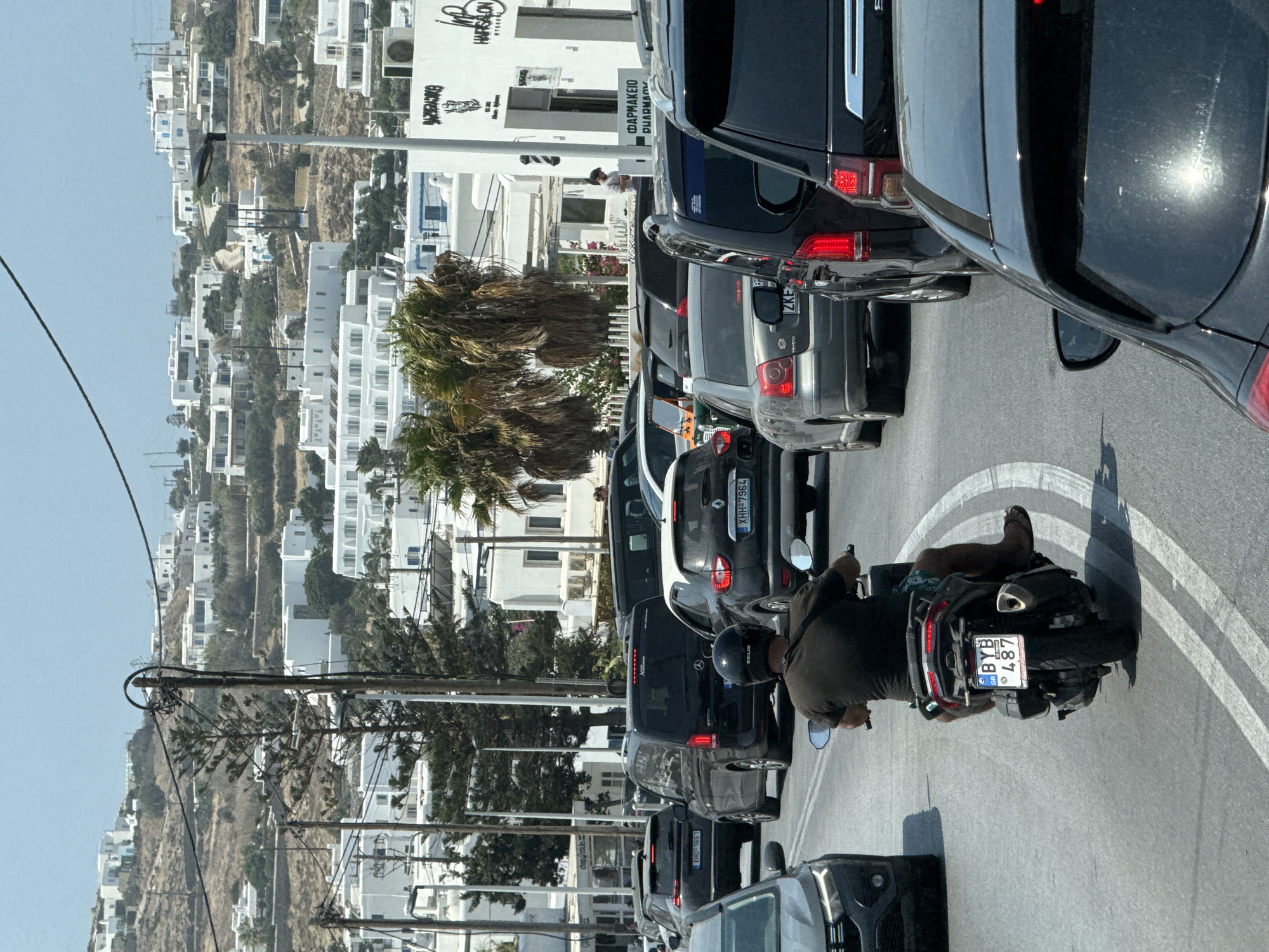 The image depicts a street scene in Ornos, Greece, with a traffic jam. A person on a scooter is in the foreground, moving away from the viewer. The scooter has a license plate reading "BYB 487". Several cars and vans are visible in the traffic, including a black Mercedes-Benz van. In the background, whitewashed buildings characteristic of Greek island architecture are seen on a hillside, with palm trees interspersed among them. The sky is clear and blue.