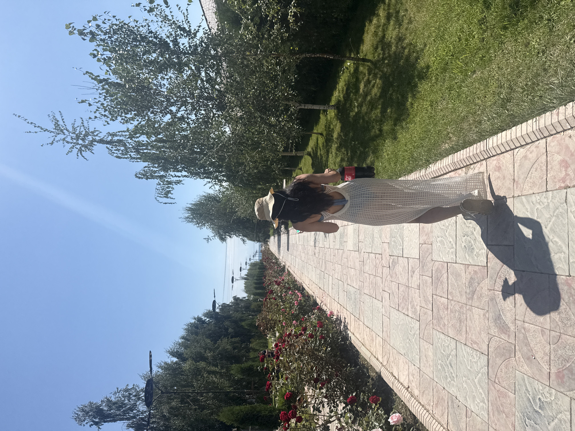 In Sary-Oy, Kyrgyzstan, a person walks down a tiled pathway lined with rose bushes on the left and trees on the right. The person, viewed from behind, wears a wide-brimmed hat, a white patterned dress, and is holding a bottle of Coca-Cola. The sun casts a long shadow of the person on the pathway. The pathway leads towards a distant horizon under a clear blue sky.