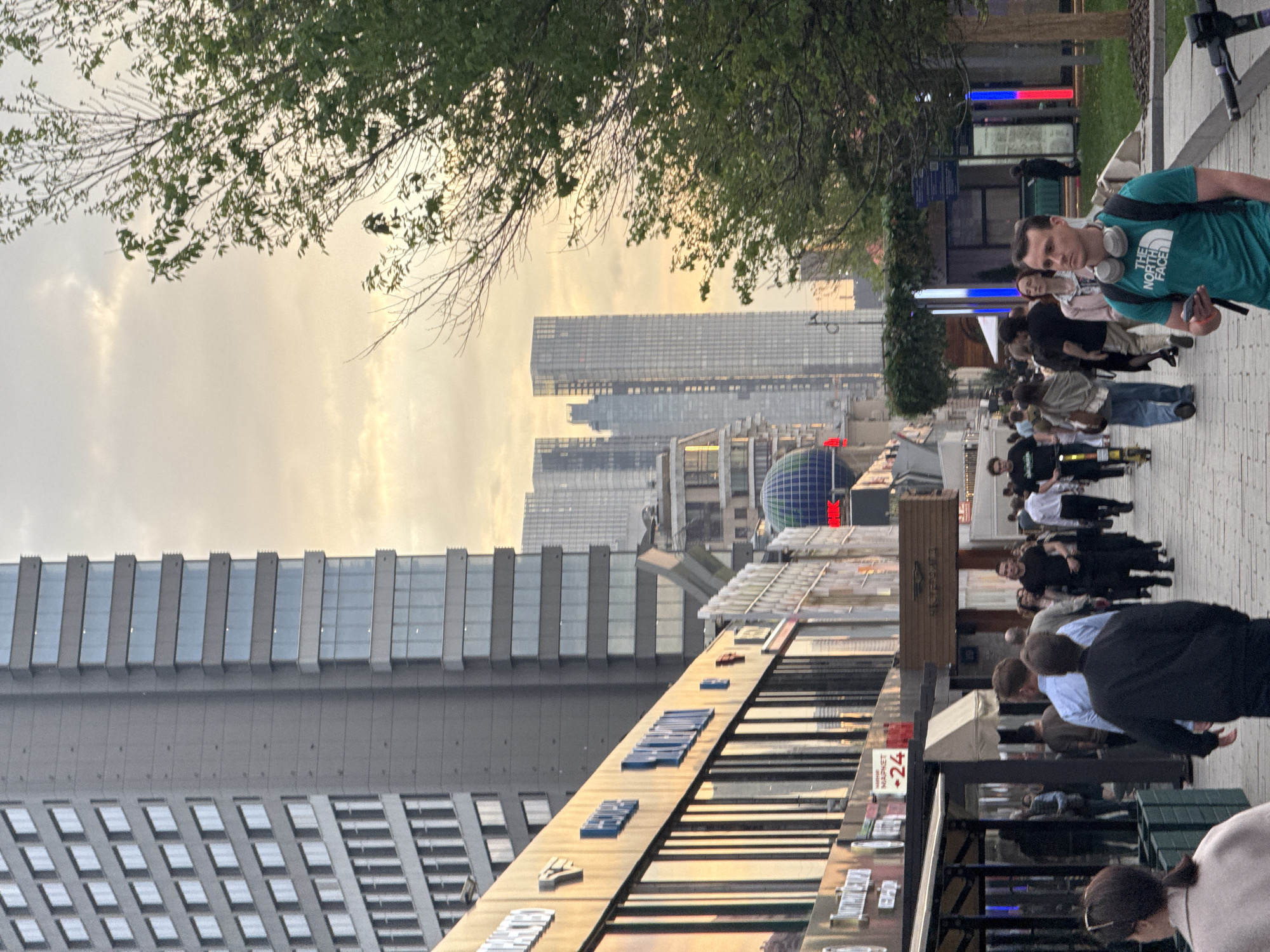 The image depicts a street scene in Moscow, Russia, with numerous pedestrians walking on a brick sidewalk. A man with a green North Face shirt and headphones is in the foreground, holding a mobile phone. Shops and outdoor seating are visible on one side of the street. Several high-rise buildings loom in the background, including a building with a distinctive blue dome. The sky is overcast, possibly at sunset. A tree branch is visible in the upper left corner. There are numerous LED signs visible.