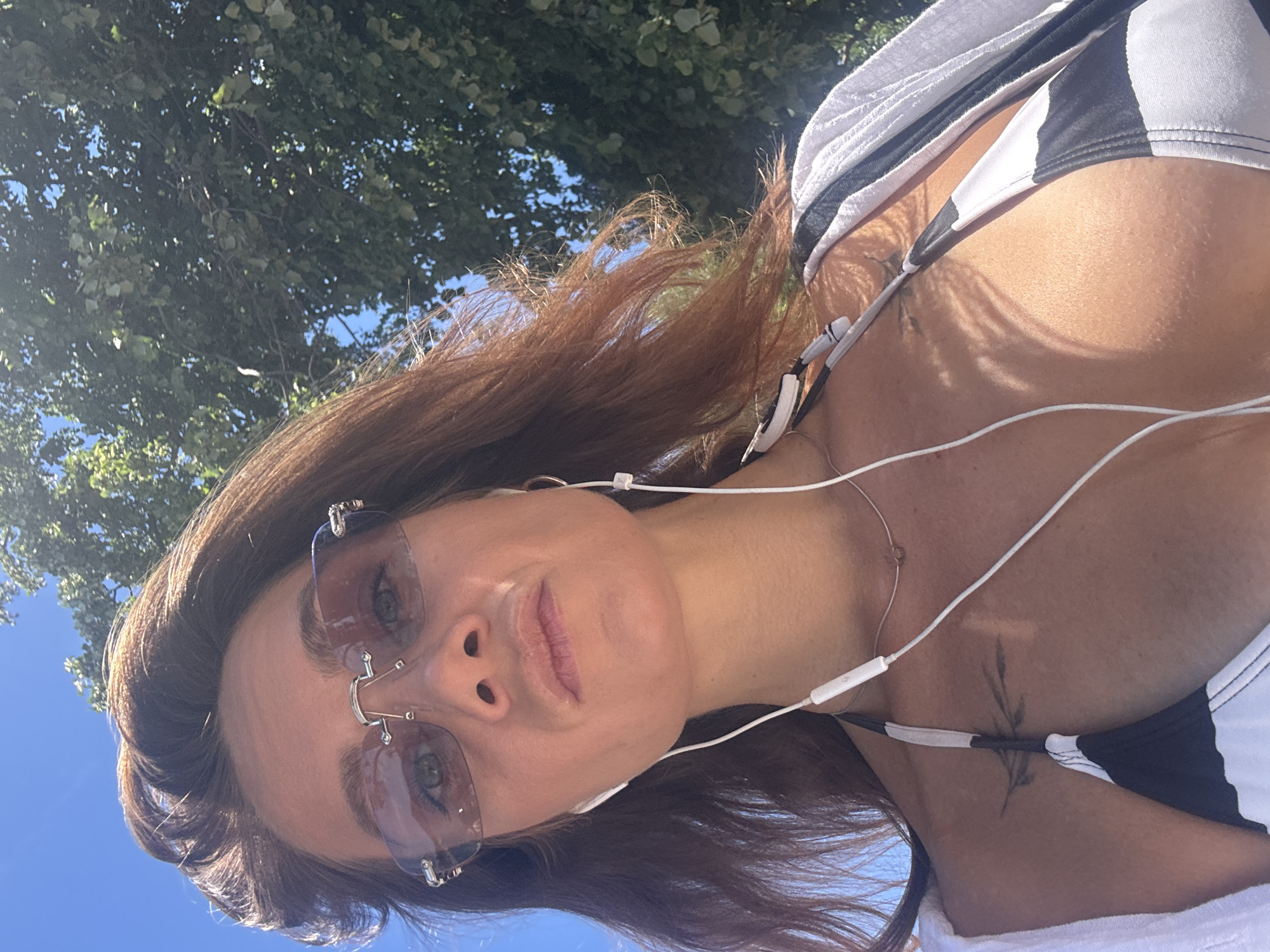 A woman, with long auburn hair and wearing sunglasses, is captured in a close-up, low-angle selfie. She is wearing a black and white striped bikini top and a light-colored open cardigan. White earphones are plugged into her ears, with the cord trailing down. She has a delicate necklace with a small pendant. A tattoo is visible on her upper chest, near her left shoulder. The background shows lush green trees and a bright blue sky, suggesting an outdoor setting in Kamenovo, Montenegro, likely on a sunny day. The lighting creates dappled shadows across her chest.
