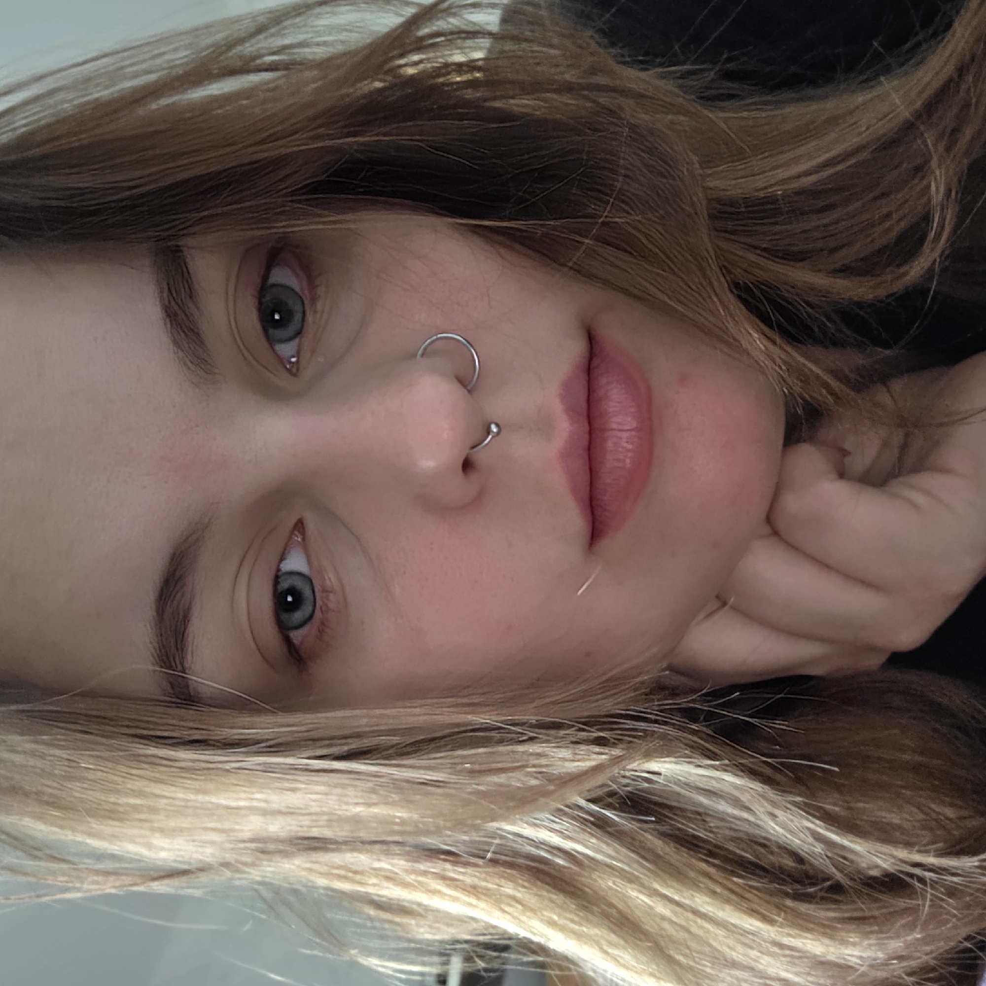 A close-up, portrait-style photograph features a person with light skin, light brown to blonde hair, and greyish-blue eyes. The person is angled so their head is tilted to the side. They have a double nose piercing, one in the upper nostril and another below. Their hand supports the side of their face. The background is dark and out of focus. The location and city are unknown.