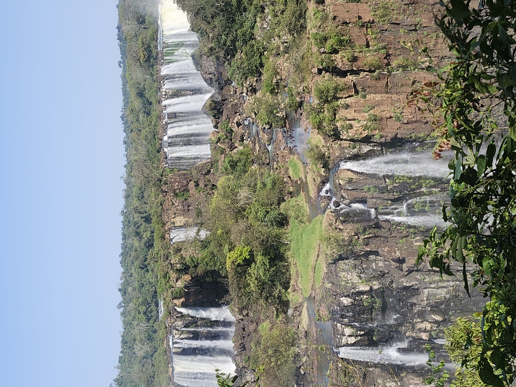 The image depicts a scenic view of Iguazu Falls, located on the border between Brazil and Argentina in Foz do Iguaçu, Brazil. The photograph showcases a wide expanse of waterfalls cascading down multiple tiers of rocky cliffs, surrounded by lush green vegetation. The sky above is a clear, bright blue. In the foreground, some foliage partially obscures the view. The waterfalls are the dominant feature, with numerous streams of water falling into a river or basin below. The cliffs are layered with various shades of brown and gray rock, interspersed with patches of green moss and vegetation. The overall scene is one of natural beauty and grandeur.