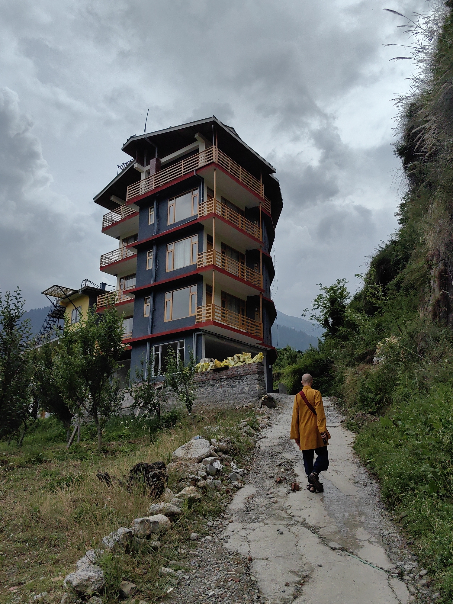 A lone, bald individual, presumably a Buddhist monk, walks away from the camera down a winding dirt and concrete path. The monk is wearing a saffron-colored robe draped over dark pants and sandals. The path is flanked by lush green vegetation and a steep, rocky hillside on the right. To the left, a multi-story building with dark blue and red accents stands prominently against a cloudy, overcast sky. The building has multiple balconies with wooden railings. In the background, hazy mountains are visible. The scene appears to be in a rural or mountainous area, consistent with Vashisht, India, known for its hot springs and proximity to the Himalayas.