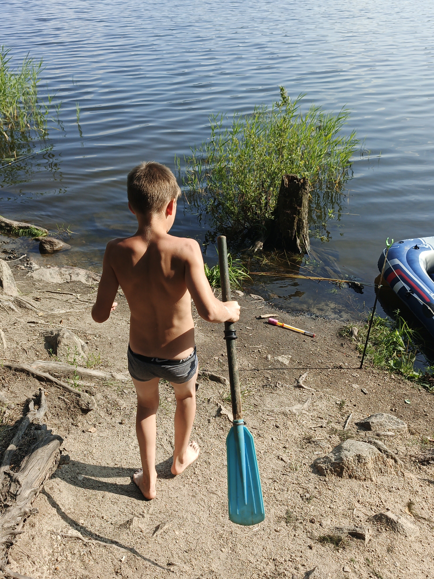 A young boy, facing away from the camera, walks barefoot on a sandy shore towards the rippling water of a lake. He is shirtless and wearing dark swim trunks, and he carries a long, blue-bladed oar. The sun casts long shadows on the ground, indicating it is likely late afternoon.
The setting is a natural lakefront, with dry driftwood and rocks scattered on the sand. Green reeds and plants grow along the water's edge, and a weathered wooden post stands in the shallow water. To the right, a deflated blue and white inflatable boat rests on the shore, with a fishing rod propped up nearby. The sky is a clear, bright blue, and the water appears calm with gentle waves. The overall atmosphere suggests a relaxed, summery day by the water. No text is visible in the image.