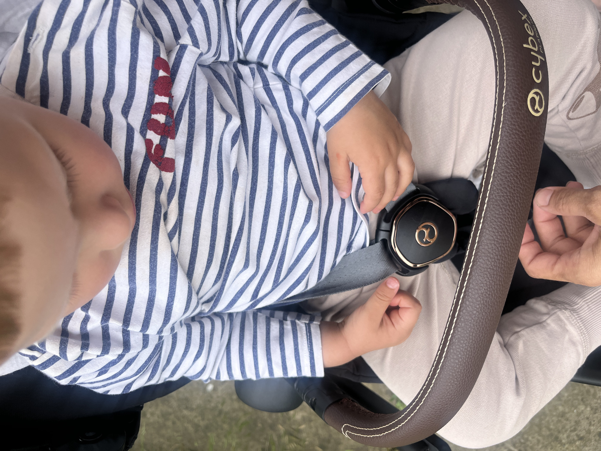A close-up shot captures a toddler in a stroller, seated and facing forward. The child wears a long-sleeved, blue and white striped shirt with the word "FUN" printed on the chest in red. A beige buckle with the Cybex logo is visible, securing the child. The stroller's brown leather handlebar, with white stitching, curves across the lower portion of the frame. A person's hand is seen adjusting something near the buckle. The background is blurred, showing hints of green and gray, suggesting an outdoor environment. The scene is set in Onokivtsi, Ukraine.