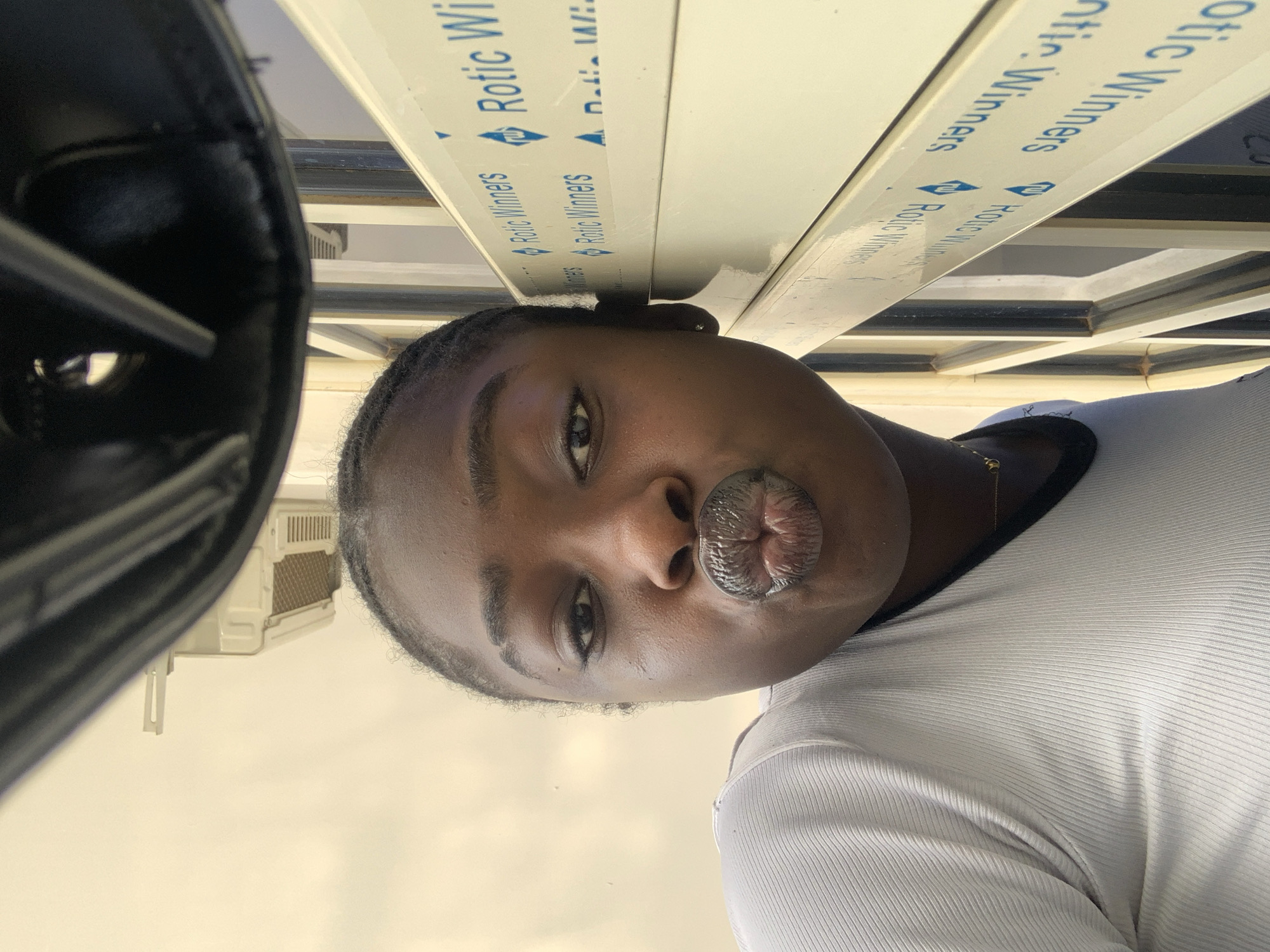 In Gura Top, Nigeria, a young woman with dark skin and braided hair is captured in a close-up selfie. She is wearing a white ribbed t-shirt with a black trim around the neckline and a delicate gold chain. Her lips are pursed in a kissing gesture, and her eyes are looking directly at the camera. The background reveals a window frame with white horizontal panels, some of which have blue text that reads "Rotic Winners" with a small diamond-shaped logo. An air conditioning unit is partially visible on the wall behind her, and above, a dark, curved object, possibly part of a structure or fixture, is present. The lighting suggests an outdoor or well-lit indoor setting.