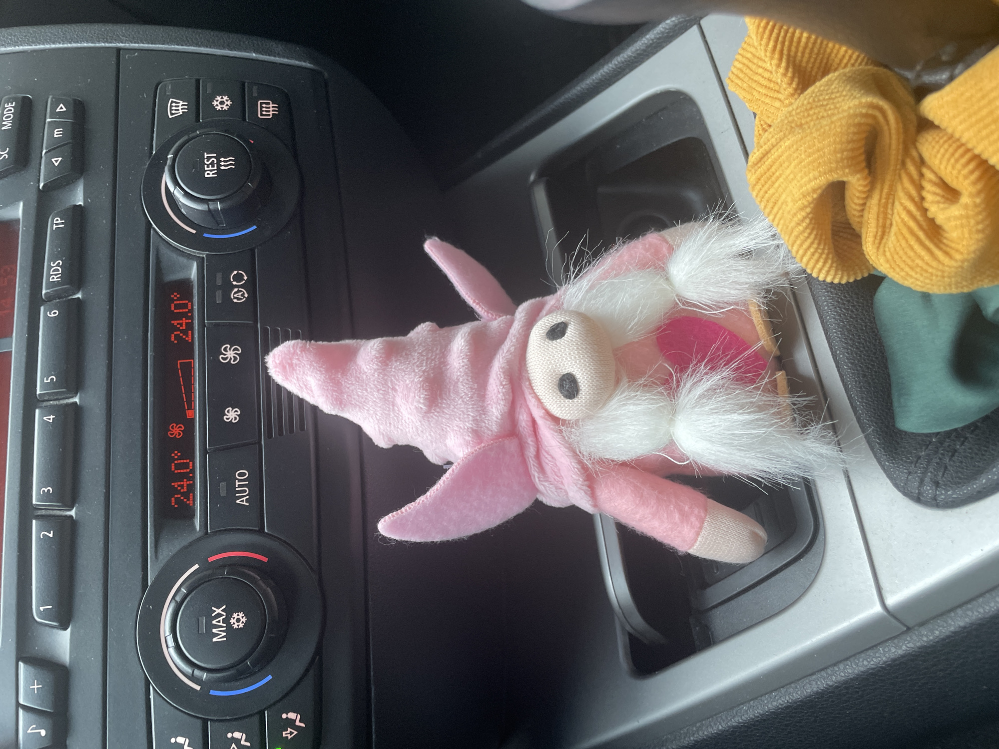 The image shows the interior of a car. A pink plush toy with a white beard and a pink hat, possibly a gnome or a similar character, is placed in the car's cup holder. Next to the cup holder, there is a yellow and green fabric item. To the left of the cup holder, there are the car's climate control buttons and display, which reads "24.0°". The "REST" and "MAX" buttons are visible. The image does not provide any information about the location in Sofia, Bulgaria, people, actions, or interactions.
