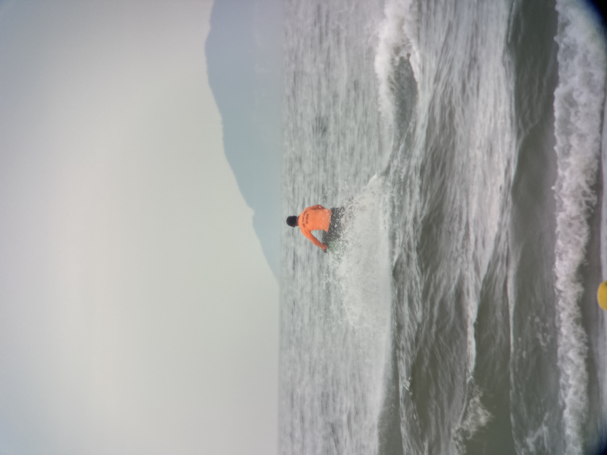 A person wearing an orange shirt is riding a jet ski on the water. The jet ski is creating a splash. In the background, there are waves and a distant mountain. The sky is overcast. The text on the orange shirt appears to be in Vietnamese. The scene is likely taking place in the waters off the coast of Đà Nẵng, Vietnam, a city known for its beaches and water sports.