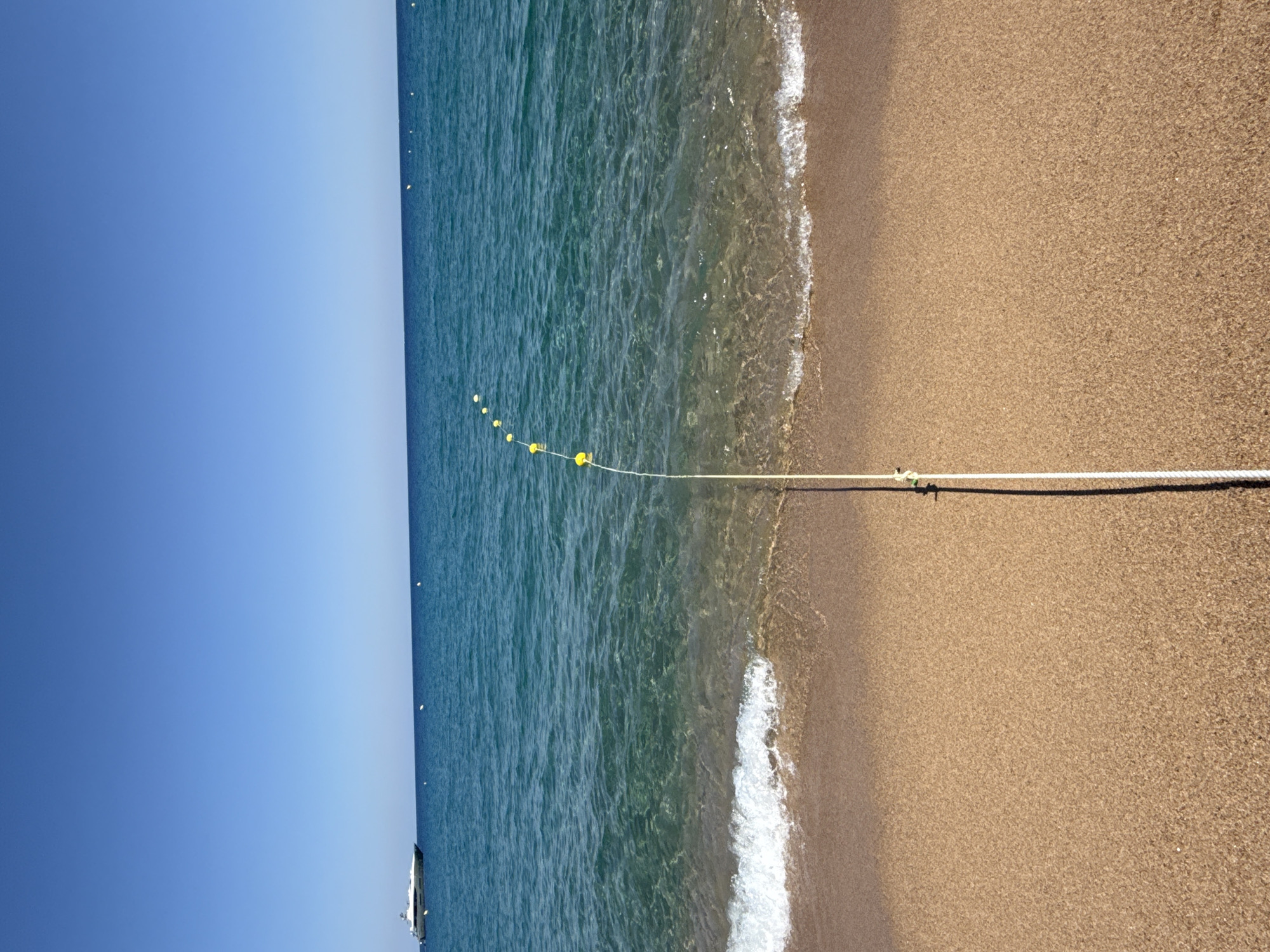 This image captures a serene beach scene in Blanes, Spain. The clear blue sky meets the tranquil turquoise waters of the Mediterranean Sea. A thin white rope, strung with small yellow buoys, stretches across the foreground, demarcating an area of the water. The sandy beach is smooth and dry, with gentle waves lapping at its edge, leaving a frothy white line of surf.
In the distance, a sleek white yacht can be seen sailing on the calm sea. The sun appears to be high in the sky, suggesting it is likely midday or early afternoon. The overall atmosphere is one of peacefulness and relaxation, typical of a sunny day at the coast. No text is visible in the image.