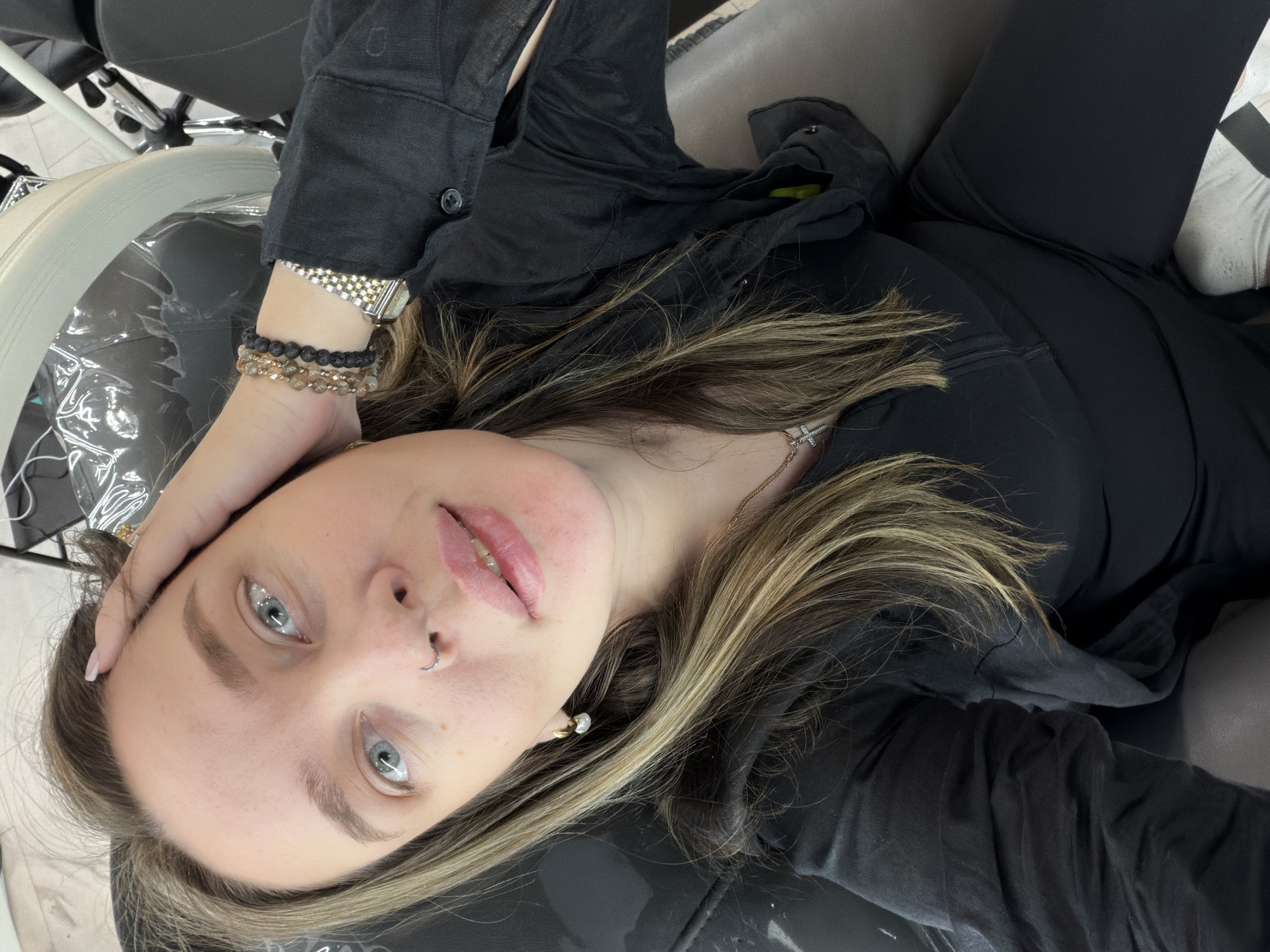 The image is a selfie-style photograph of a fair-skinned individual with light blue eyes and blonde hair with darker roots. They are wearing a black button-down shirt and black pants. A silver nose ring is visible in their left nostril and a small gold earring is visible in their right ear. The person is wearing a watch and several bracelets on their left wrist. They are seated in a black chair and their left hand rests on their forehead.
The environment appears to be indoors, possibly in a salon or a similar establishment, as suggested by the metallic equipment with plastic covers visible in the periphery on the left side.
I cannot confidently pinpoint the exact location in Moscow, Russia, as there are no discernable landmarks or details specific enough to determine the geographic location.